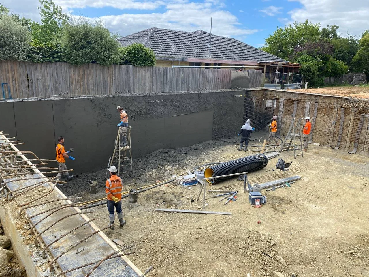 Construction workers applying concrete to a large wall at a construction site with building materials and tools scattered around.