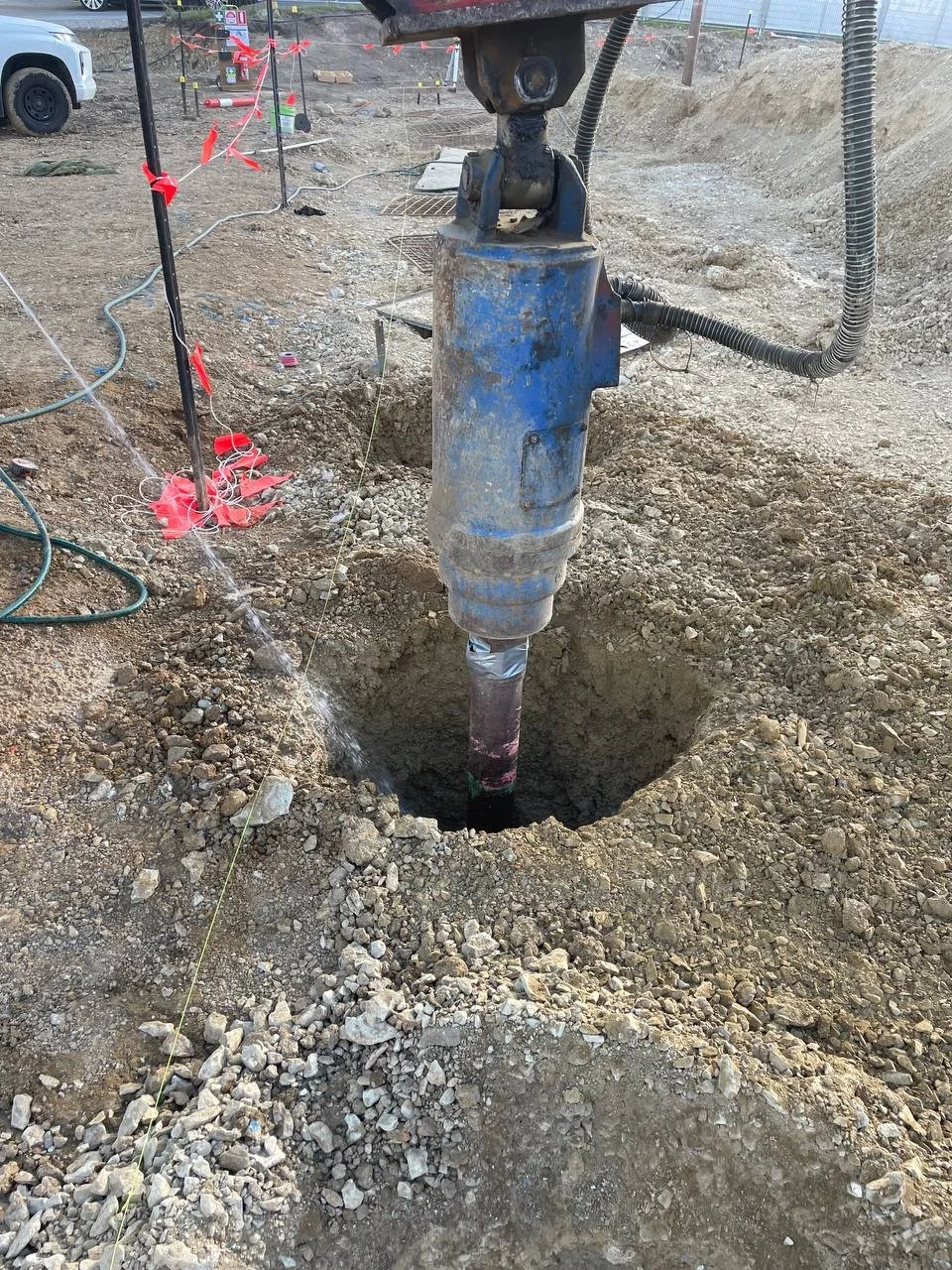 A construction site with a large hydraulic drill installing a pile into the ground, surrounded by excavated dirt, with construction and safety markings nearby.