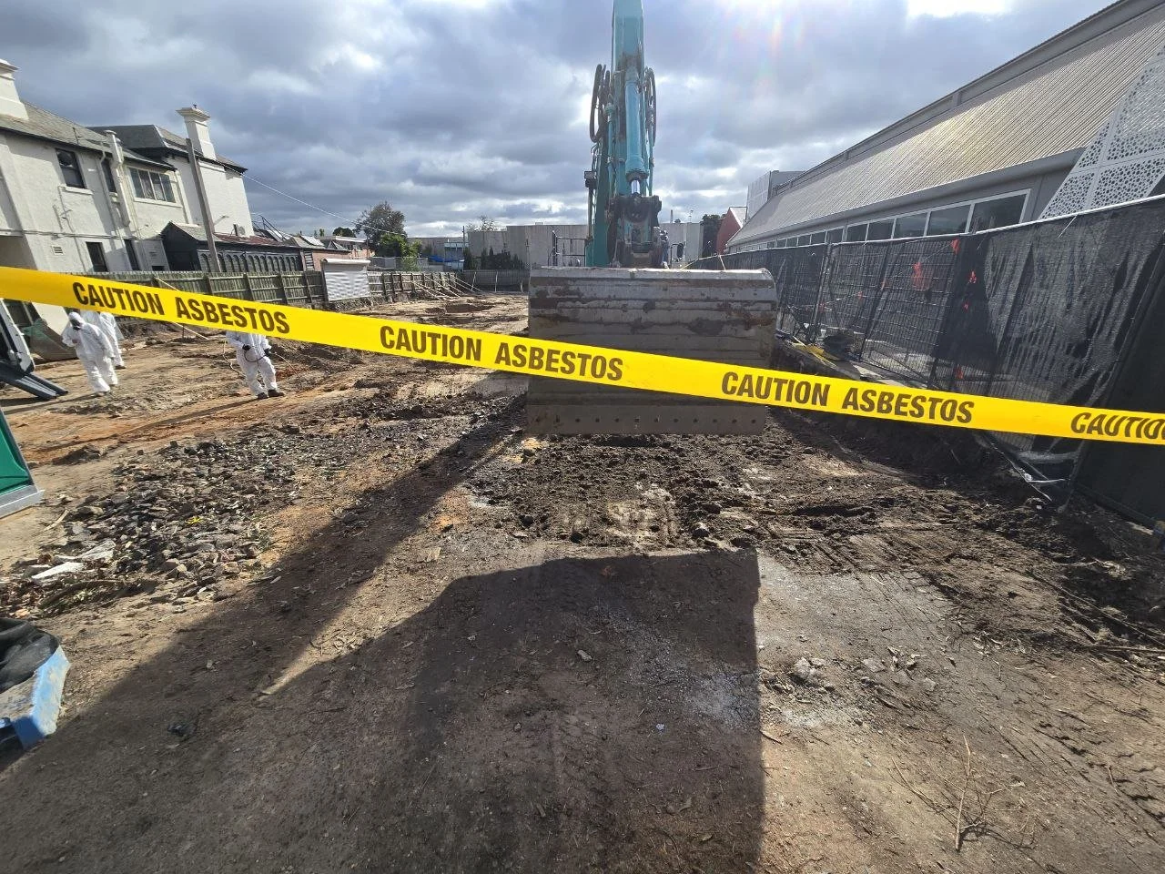 Construction site with heavy machinery and caution tape reading 'Caution Asbestos,' workers in protective gear, and partially demolished ground under cloudy skies.