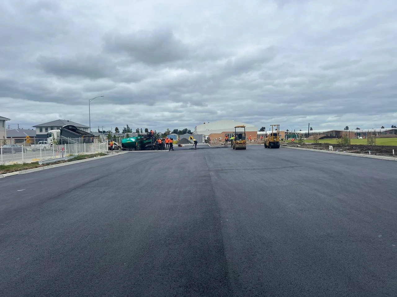 Workers paving a newly constructed road, with construction equipment and buildings in the background, under cloudy skies.