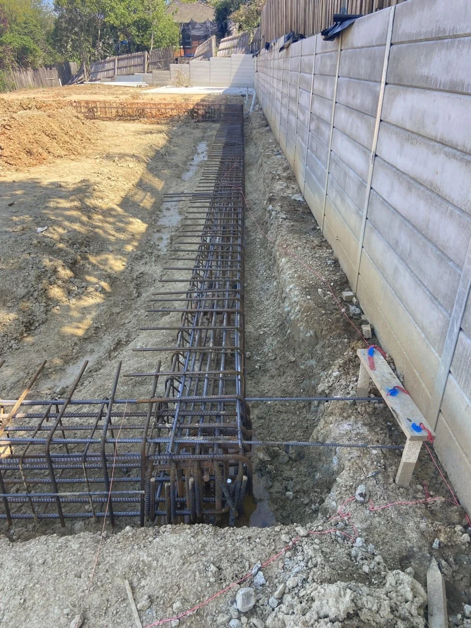Construction site with rebar reinforcement for concrete foundation along a trench, next to a wooden fence and retaining wall.
