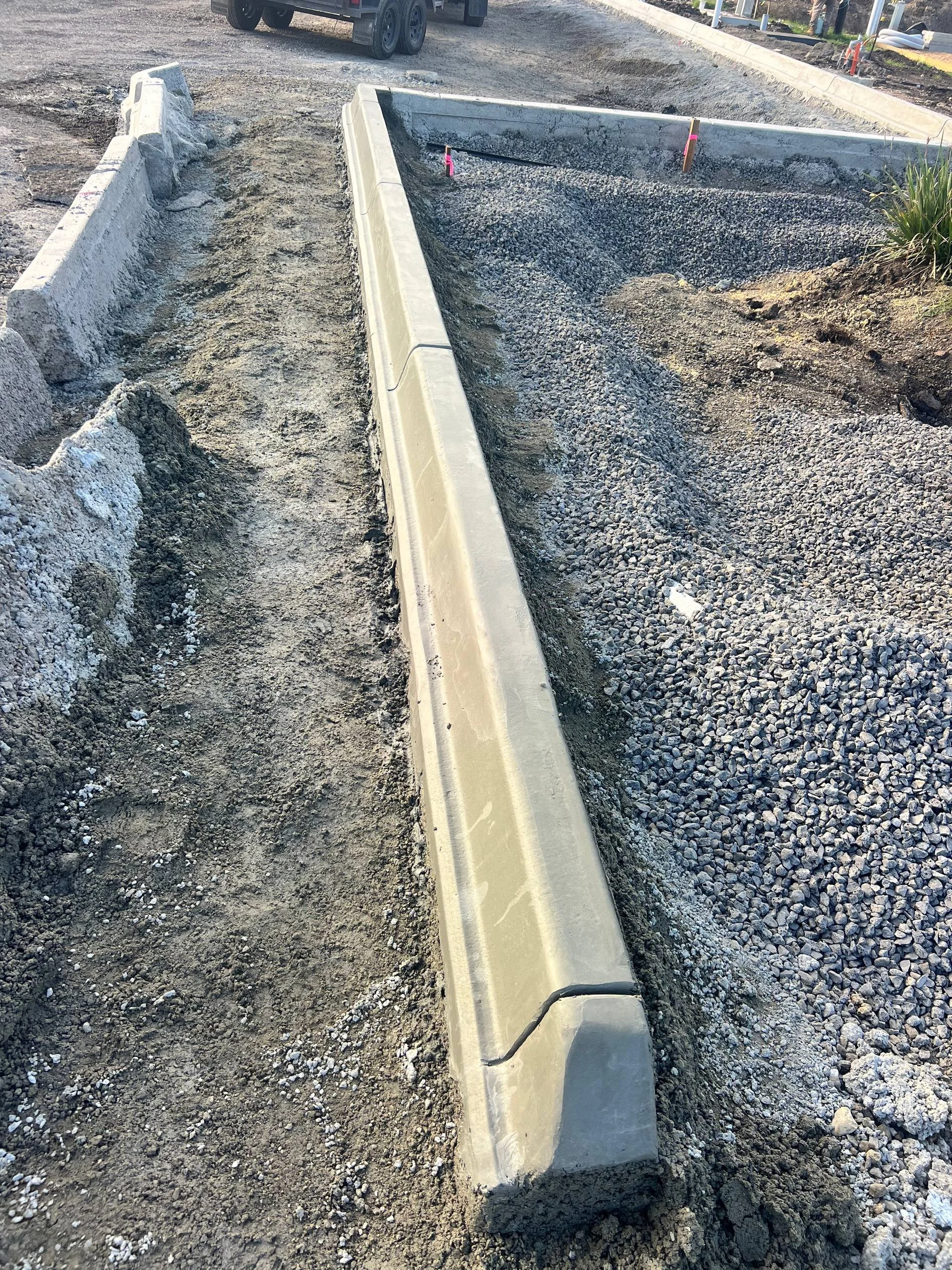 Construction site showing a concrete curb installed on a gravel surface with dirt and soil on both sides, and construction markings visible. A vehicle is parked in the background.