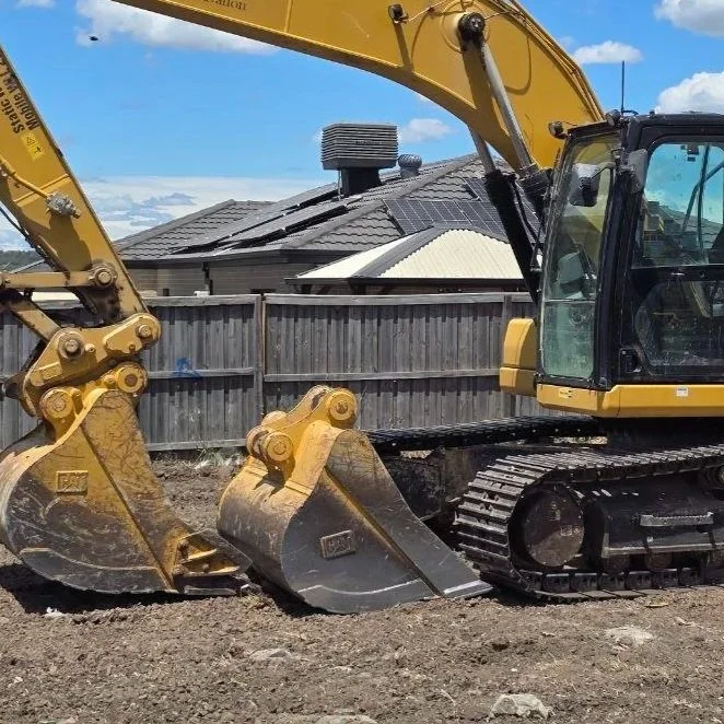 Close-up of a yellow excavator on a construction site, with a residential house and cloudy blue sky in the background.