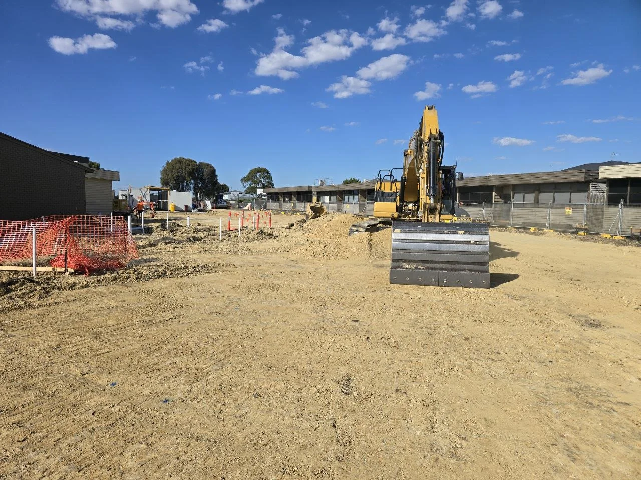 Construction site with a yellow excavator on bare dirt, orange safety fencing, and buildings in the background under a blue sky with scattered clouds.