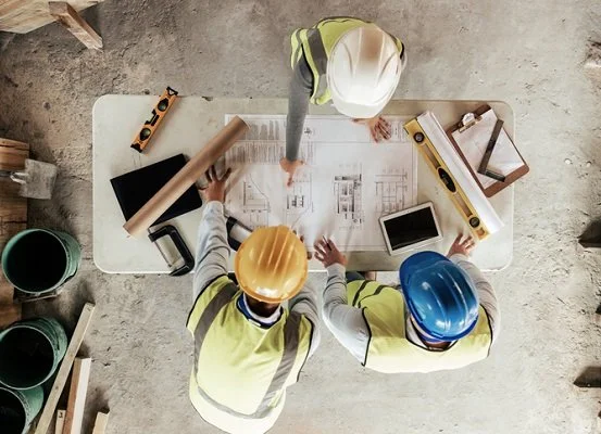 Three construction workers reviewing blueprints at a table, wearing safety helmets and vests.