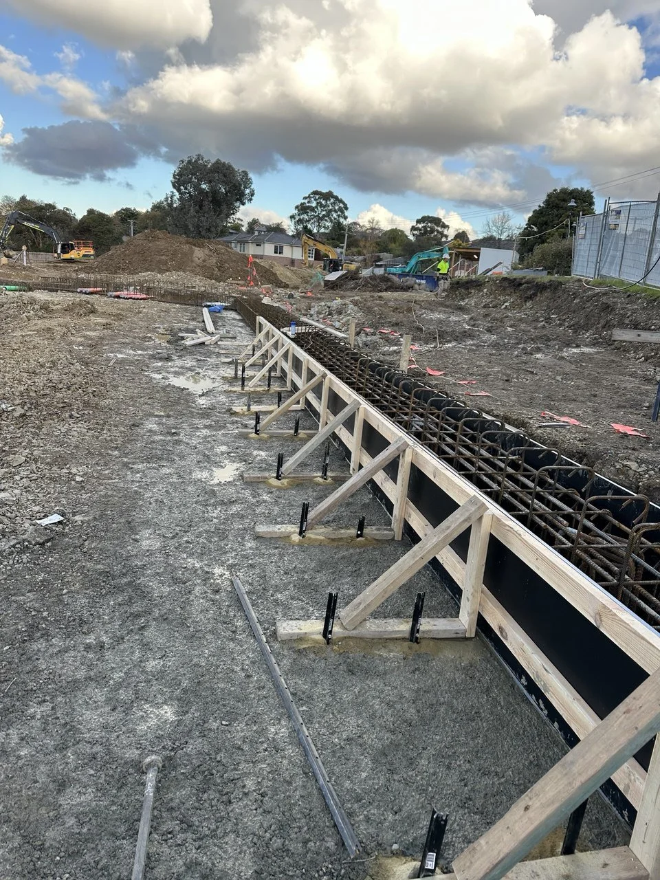 Construction site with rebar and wooden forms for a concrete foundation, heavy machinery, construction workers, and an overcast sky.