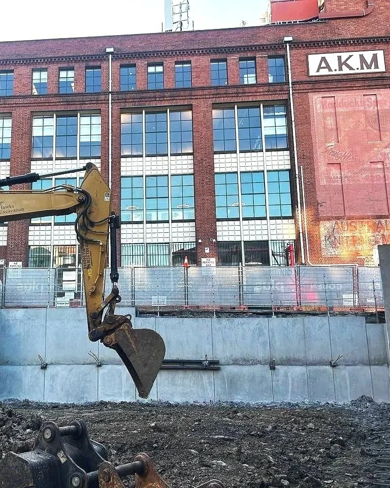 Construction site in front of a brick building with large windows, construction fencing, and excavation equipment.
