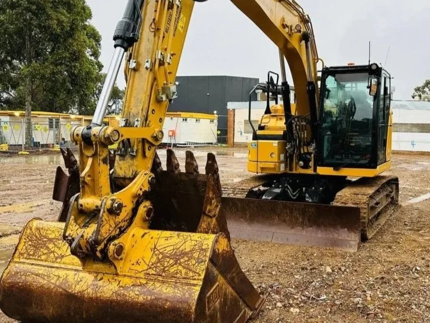 A yellow excavator on a construction site with dirt and gravel, surrounded by some fences and background buildings.