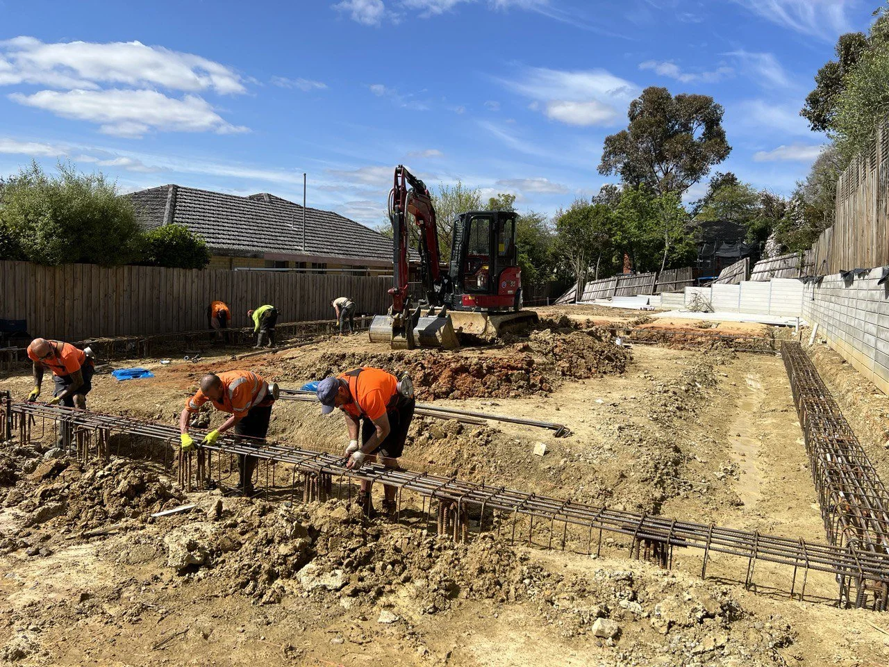 Construction workers laying rebar at a construction site with a small excavator, during a sunny day with blue sky and trees in the background.