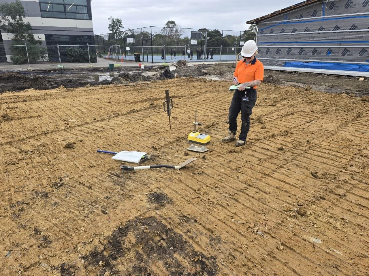 A construction worker in an orange shirt and white helmet reviews notes at a construction site with freshly tilled soil. Construction tools are on the ground, and a building with a partially covered wall and a fenced sports court are visible in the b