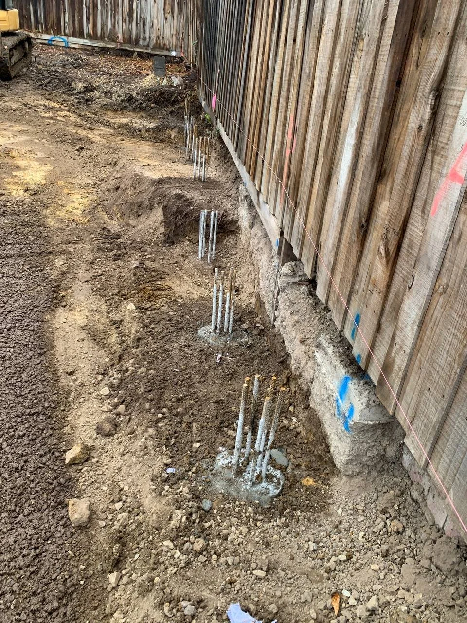 Construction site with concrete footing and exposed rebar protruding from the ground along a wooden fence, marking a foundation or wall.