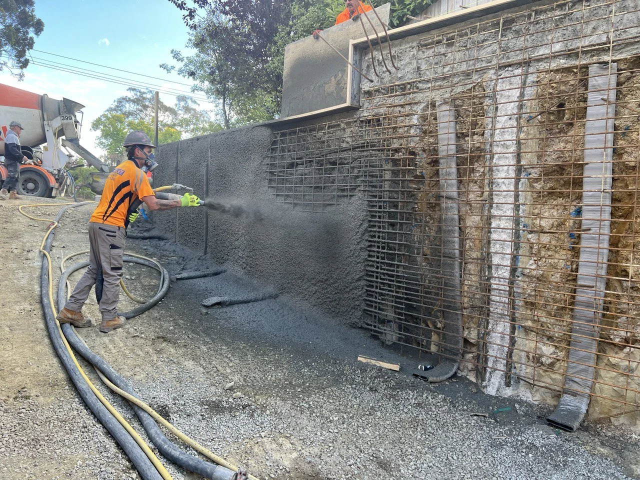Worker spraying concrete on a building foundation with rebar, wearing safety gear including helmet and gloves.