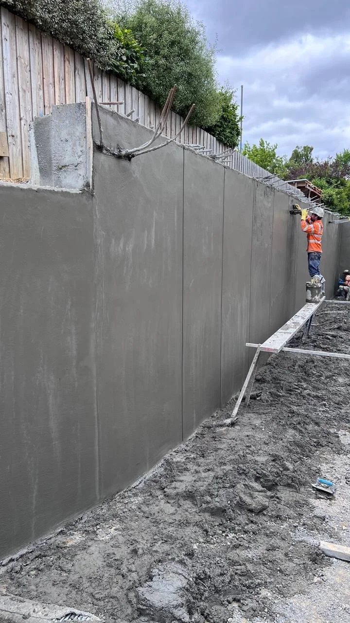 Construction workers applying gray concrete to a wall outdoors, with one worker standing on a small platform and another worker sitting nearby.