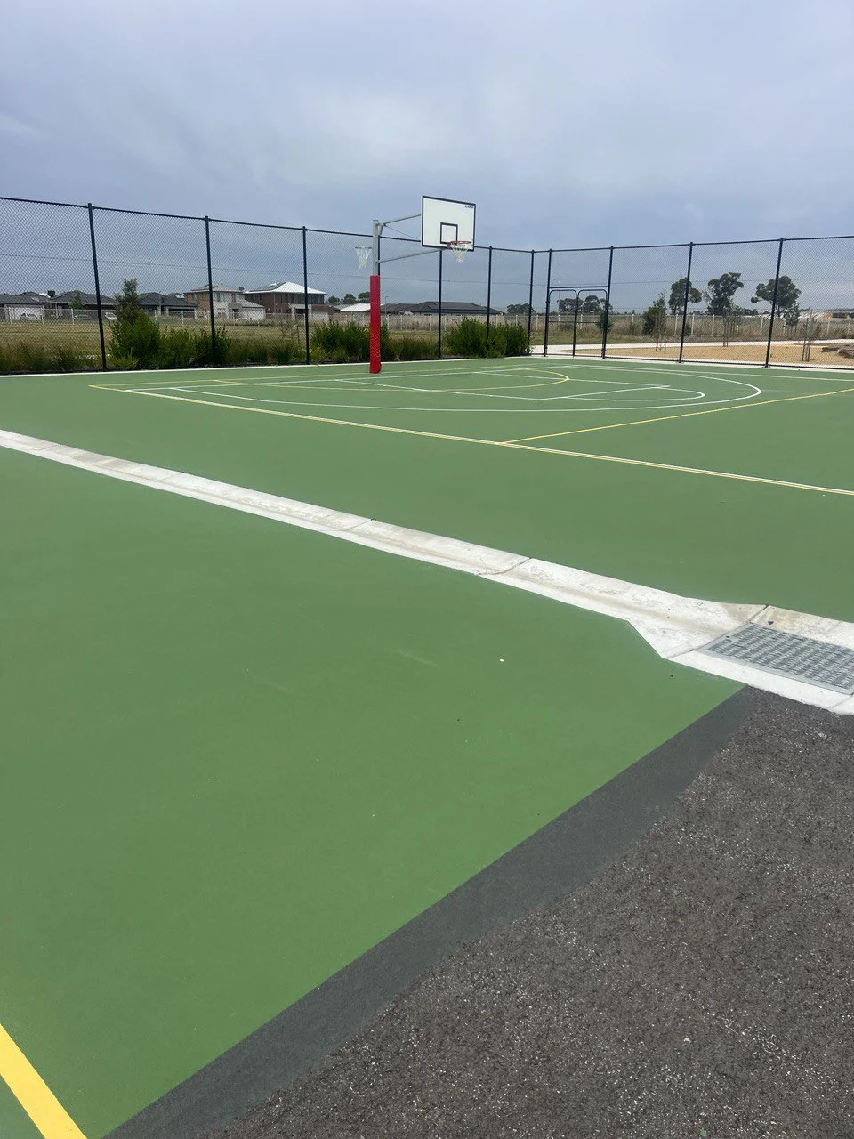 Empty outdoor basketball court with green surface, white and yellow lines, surrounded by a black chain-link fence, under overcast sky.