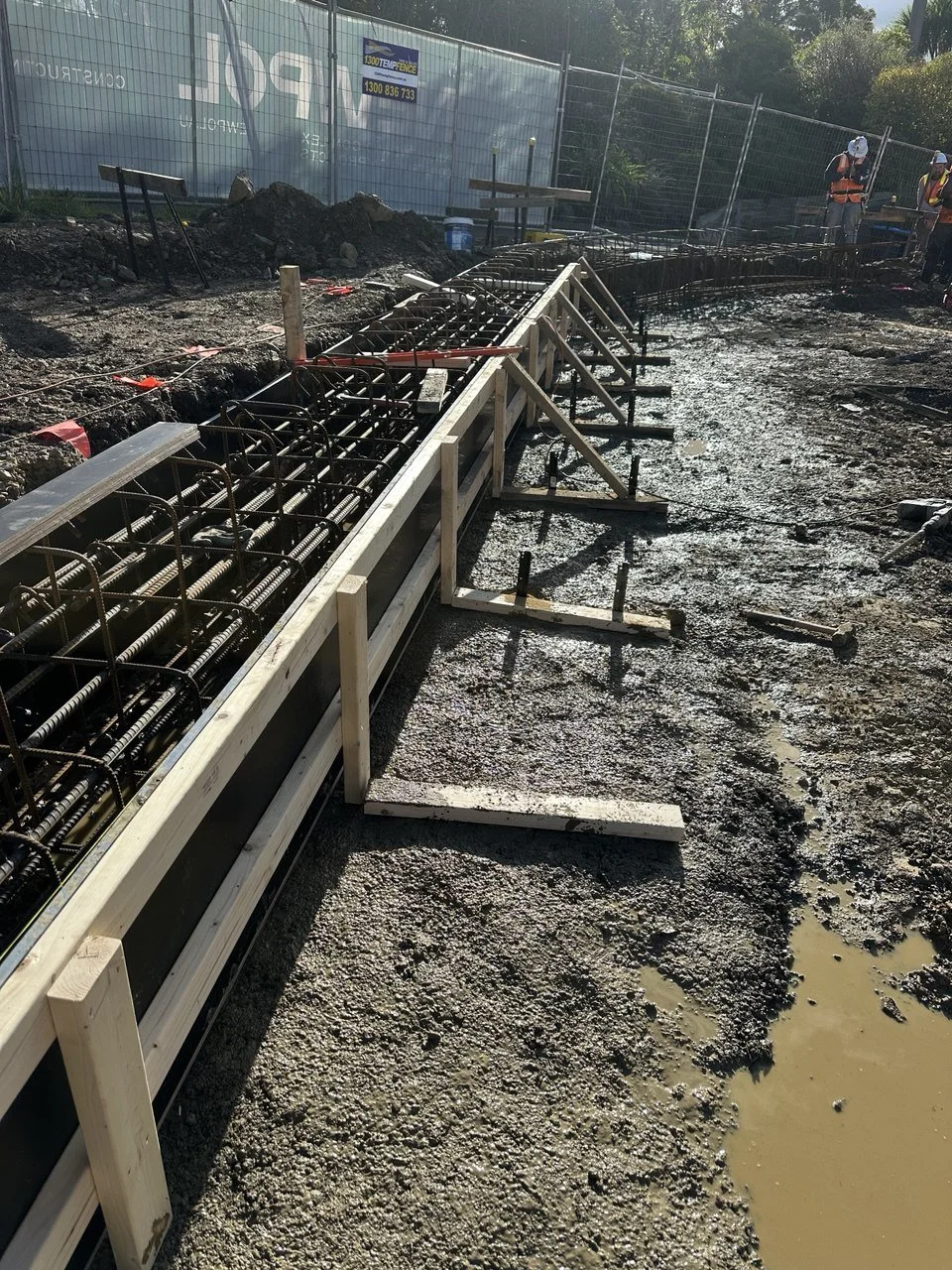 Construction site with metal rebar framework and wooden formwork for a concrete structure. Two construction workers in orange safety vests and helmets are visible in the background. The ground is muddy with some standing water.