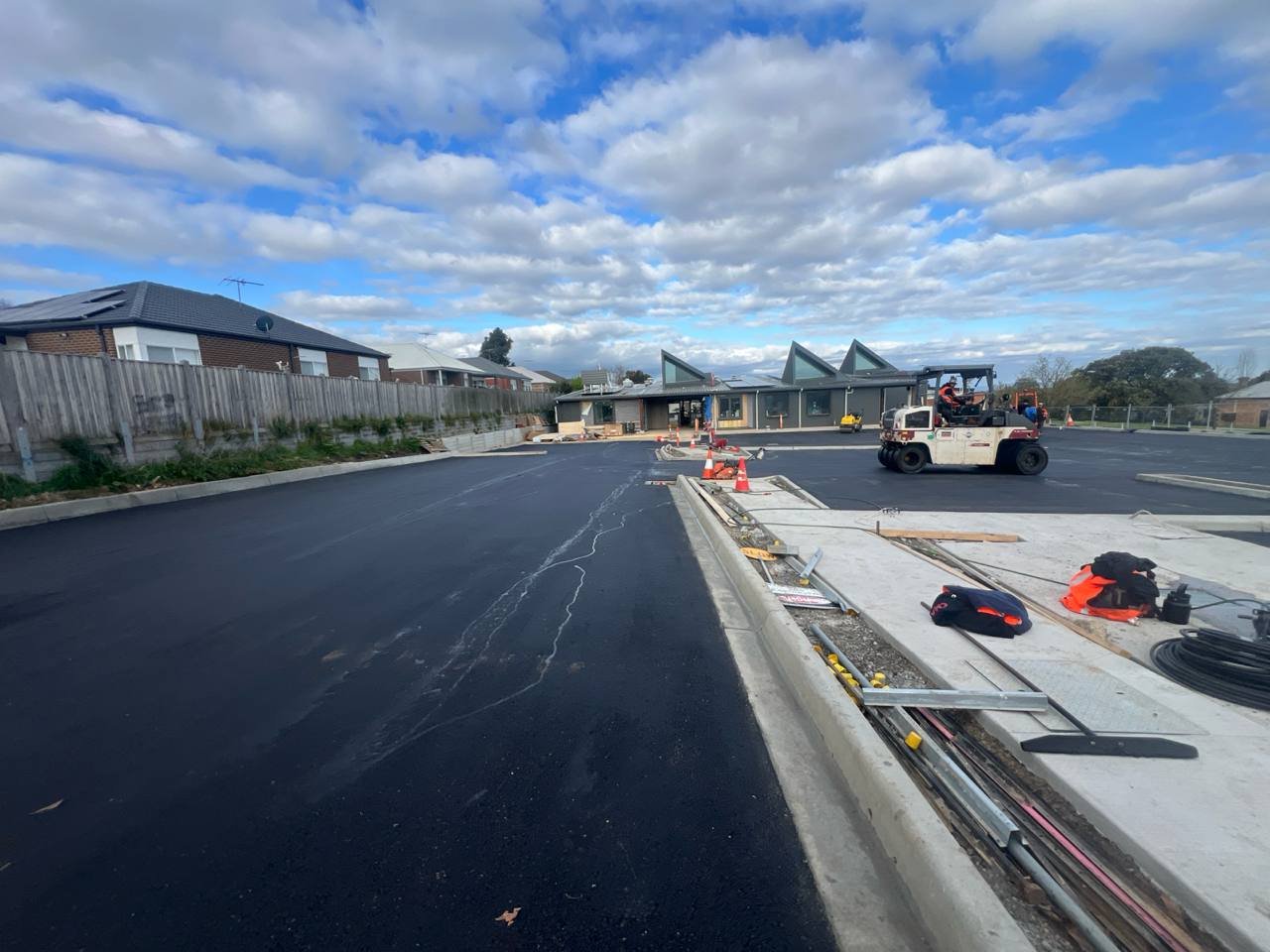 Construction site with newly paved black asphalt, construction equipment, and unfinished sidewalk.
