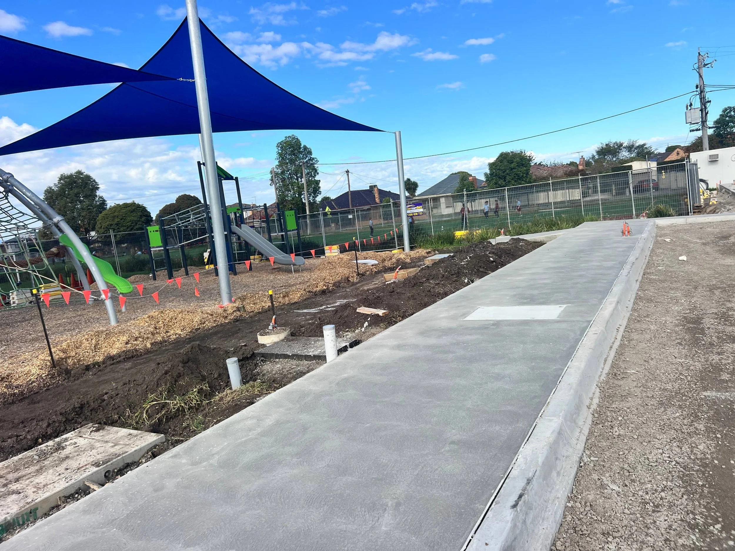 Construction site with a newly poured sidewalk, construction barriers, playground equipment with shade sails, and residential houses and trees in the background.
