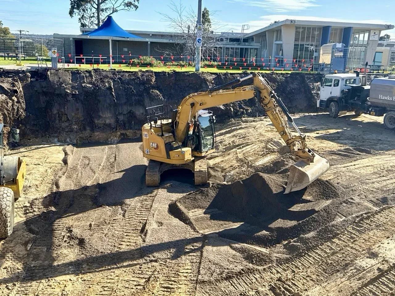 Construction site with a yellow excavator digging into the ground, trucks parked nearby, and a building with a blue roof and glass windows in the background.