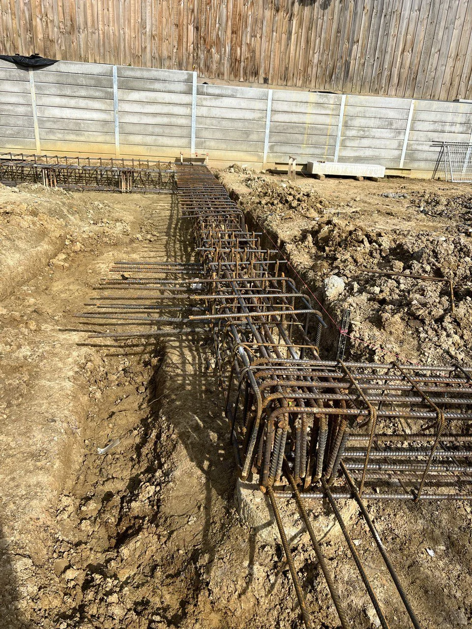 Under construction site showing exposed steel rebar reinforcement in a trench, with a wooden and metal fence in the background.
