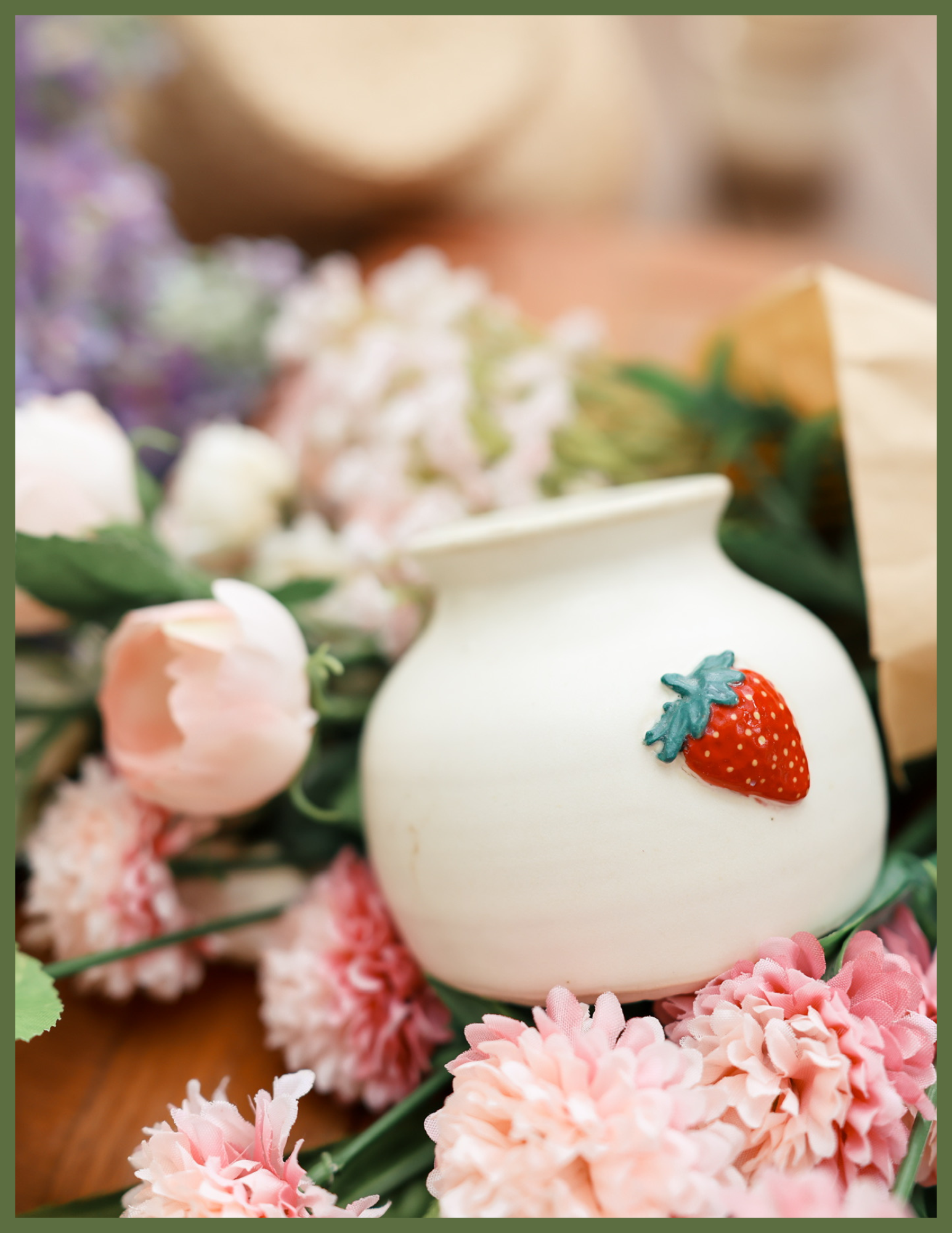 Close-up of a white, possibly ceramic, object with a strawberry decoration on it, surrounded by pink and white flowers, and some greenery.