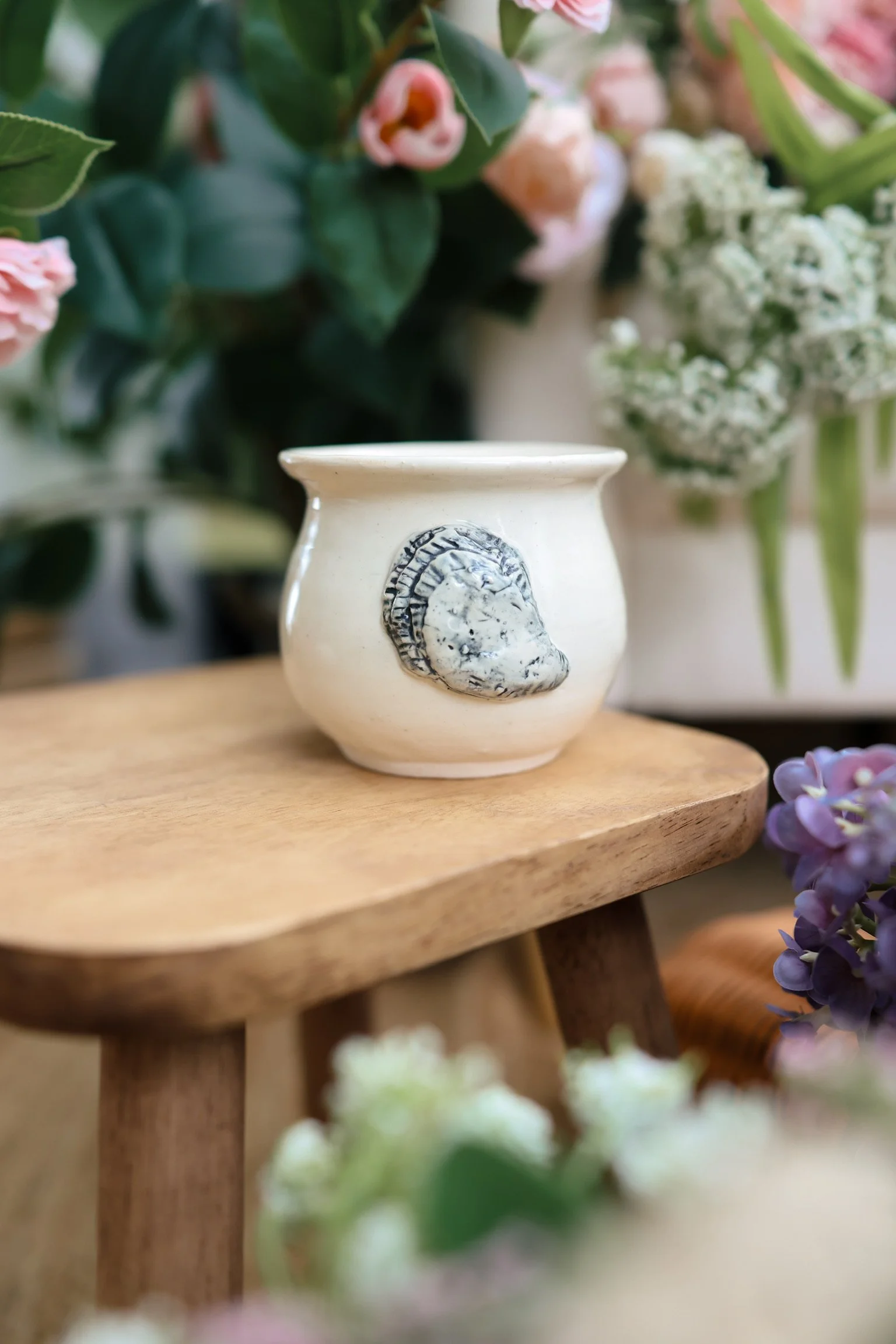 Close-up of a white decorative vase with a gray face cameo, placed on a wooden surface with a background of pink and white flowers and green leaves.