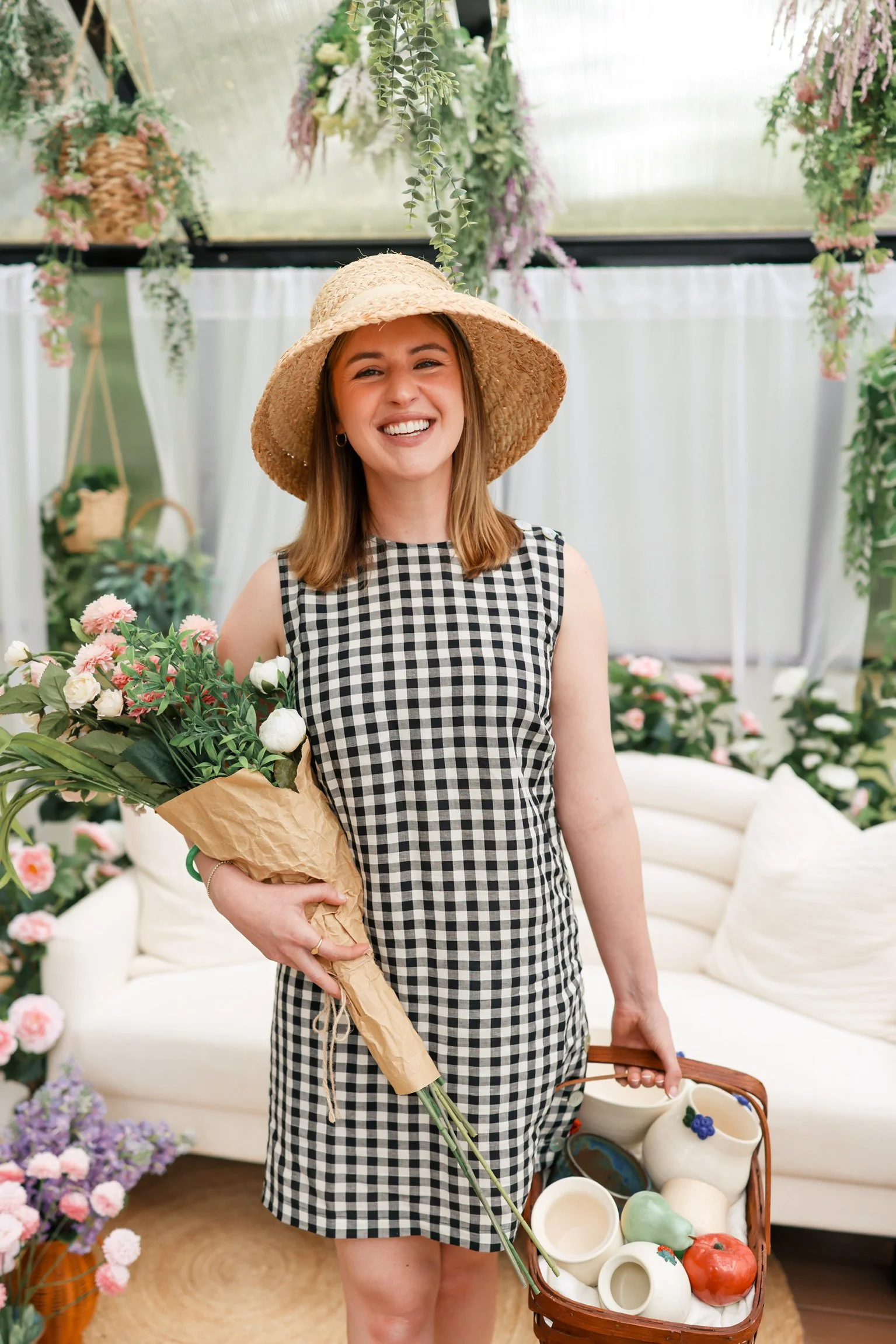 A woman in a black and white checkered dress and a straw hat holding a bouquet of flowers and a basket filled with pottery and fruits inside a floral shop or greenhouse.