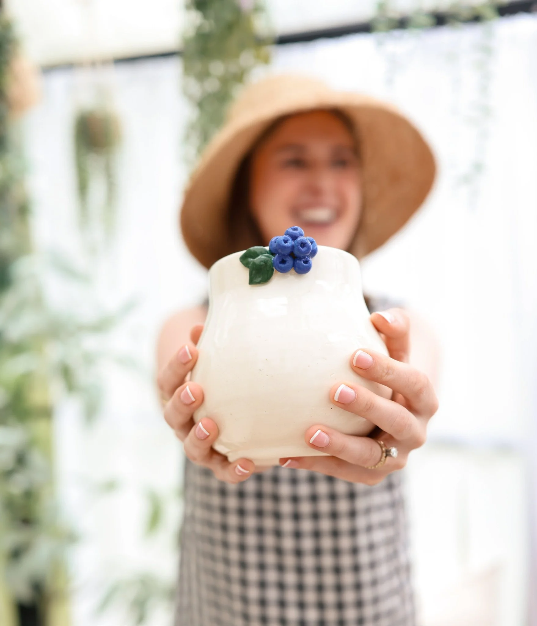 Person wearing a wide-brimmed hat, holding a small white ceramic container decorated with blue berries and green leaves, in an outdoor setting with blurred greenery and a glass window in the background.