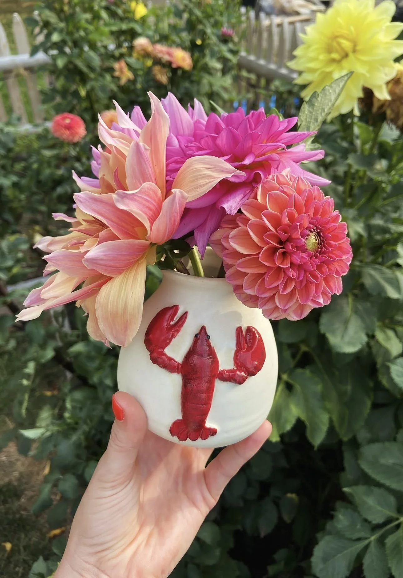 A hand holding a white vase with red lobster and lobster claw painted on it, filled with pink and peach dahlias and a yellow flower, with a background of green foliage.