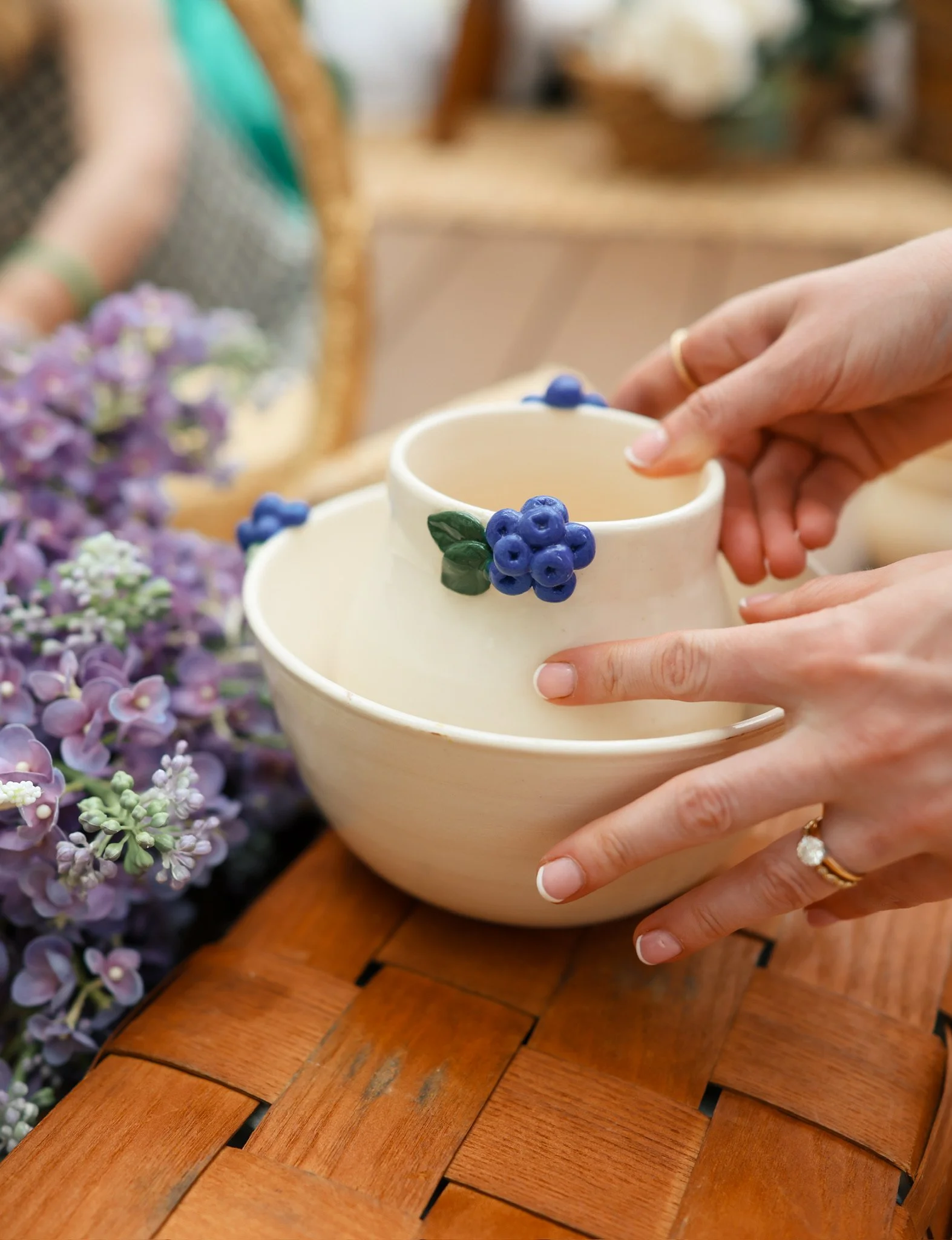 Person holding a small, white ceramic pitcher with a green leaf and blue berries decoration, sitting on a wooden table with purple hydrangea flowers nearby.