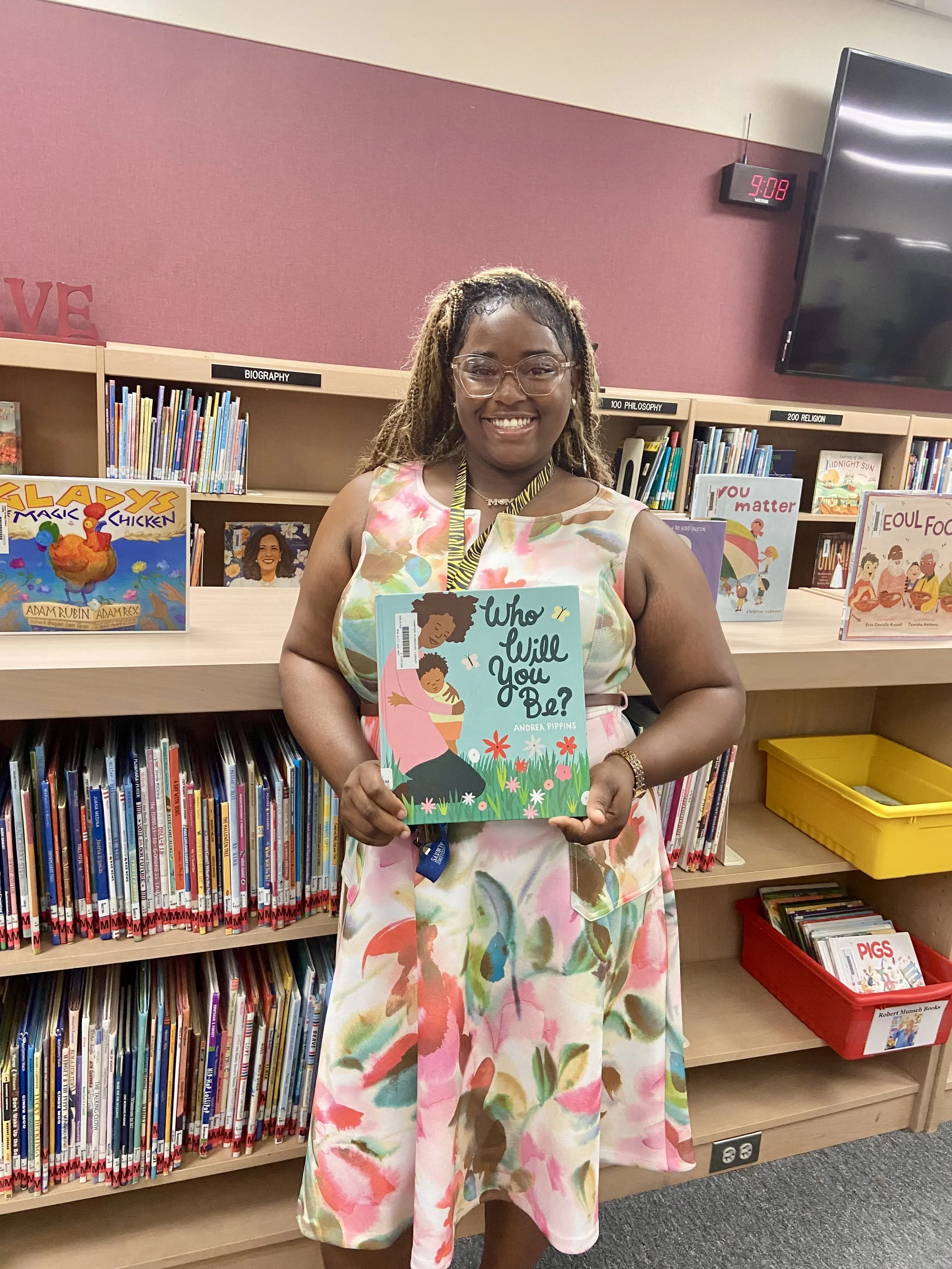 A woman with dark skin and curly hair wearing glasses and a dress with a colorful abstract pattern, standing in a library holding a children's book titled 'Who Will You Be?' by Andrea Pippins, with bookshelves filled with children's books behind her.