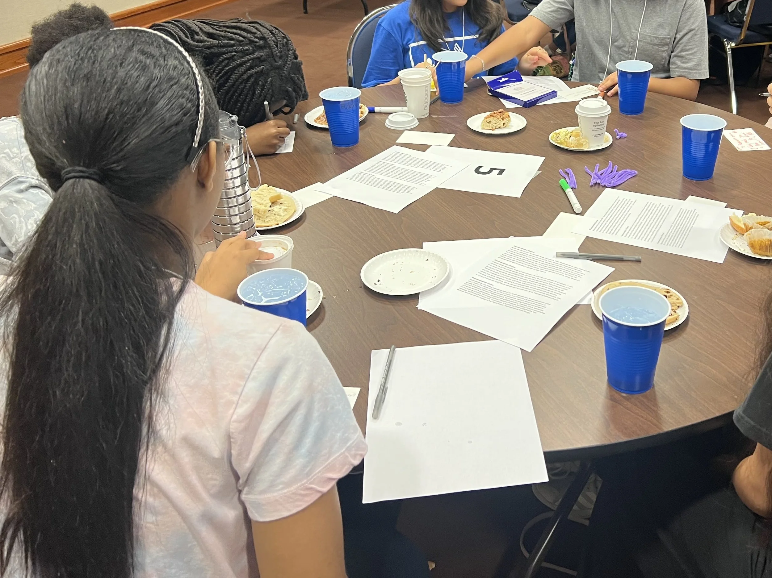 A group of people sitting around a conference table with papers, plates of food, cups, and writing utensils, engaged in a meeting or discussion.