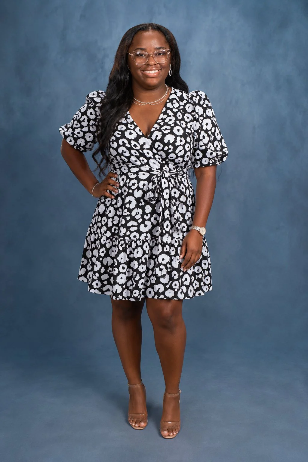 A woman standing against a blue background, smiling, wearing a black and white floral dress, clear high-heeled shoes, and glasses.