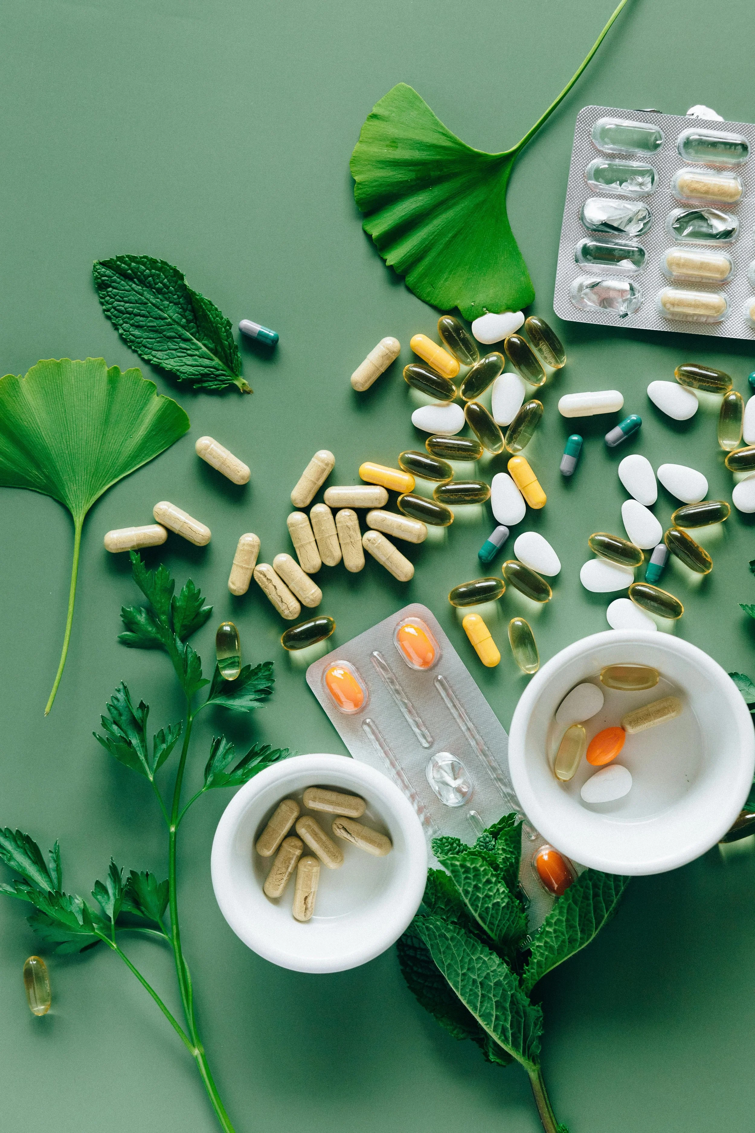 Assorted pills and capsules on a green background with green leaves, some in white bowls and blister packs.