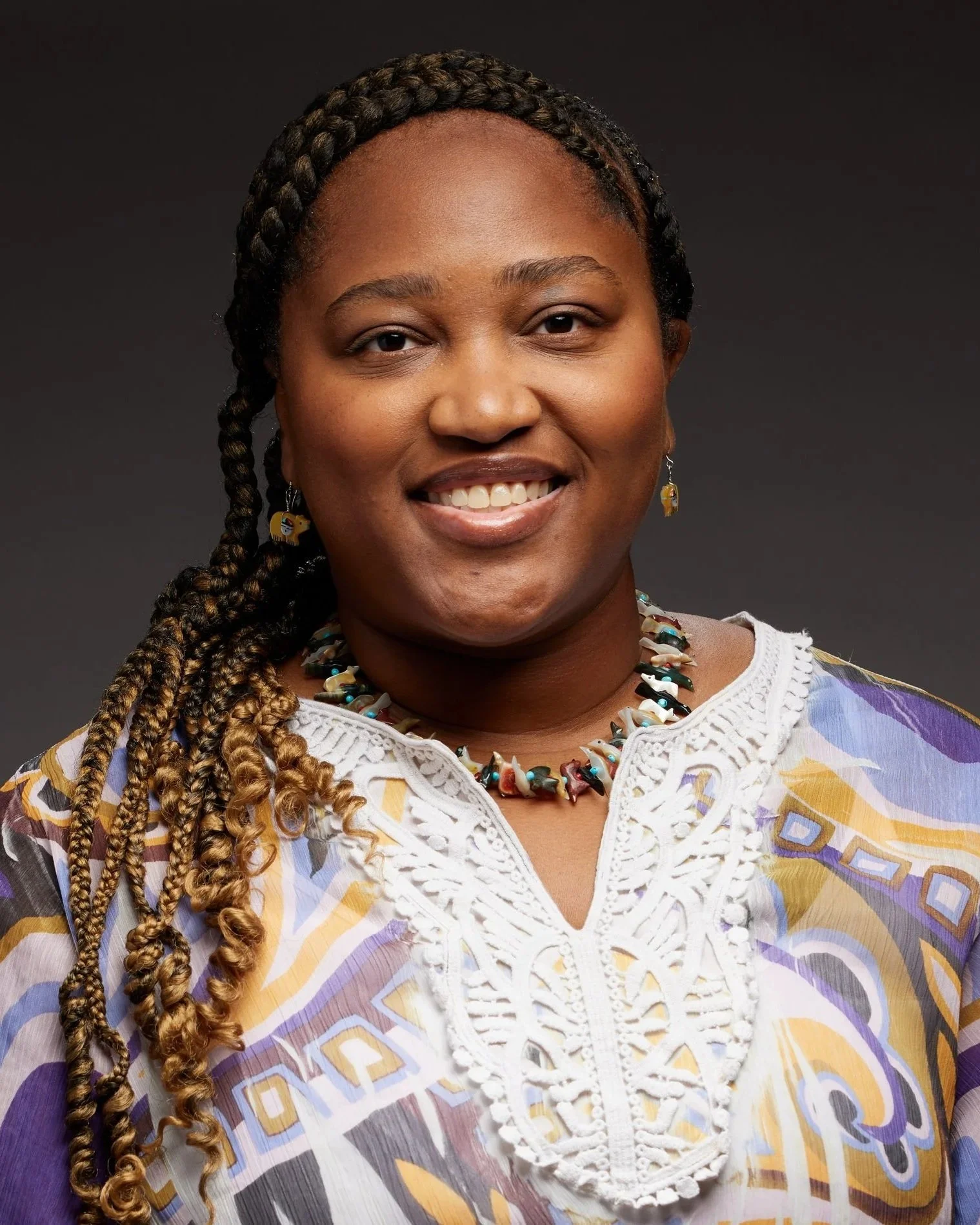 A woman with braided hair, wearing colorful earrings, a beaded necklace, and a multicolored patterned blouse with white lace detail, smiling against a dark background.