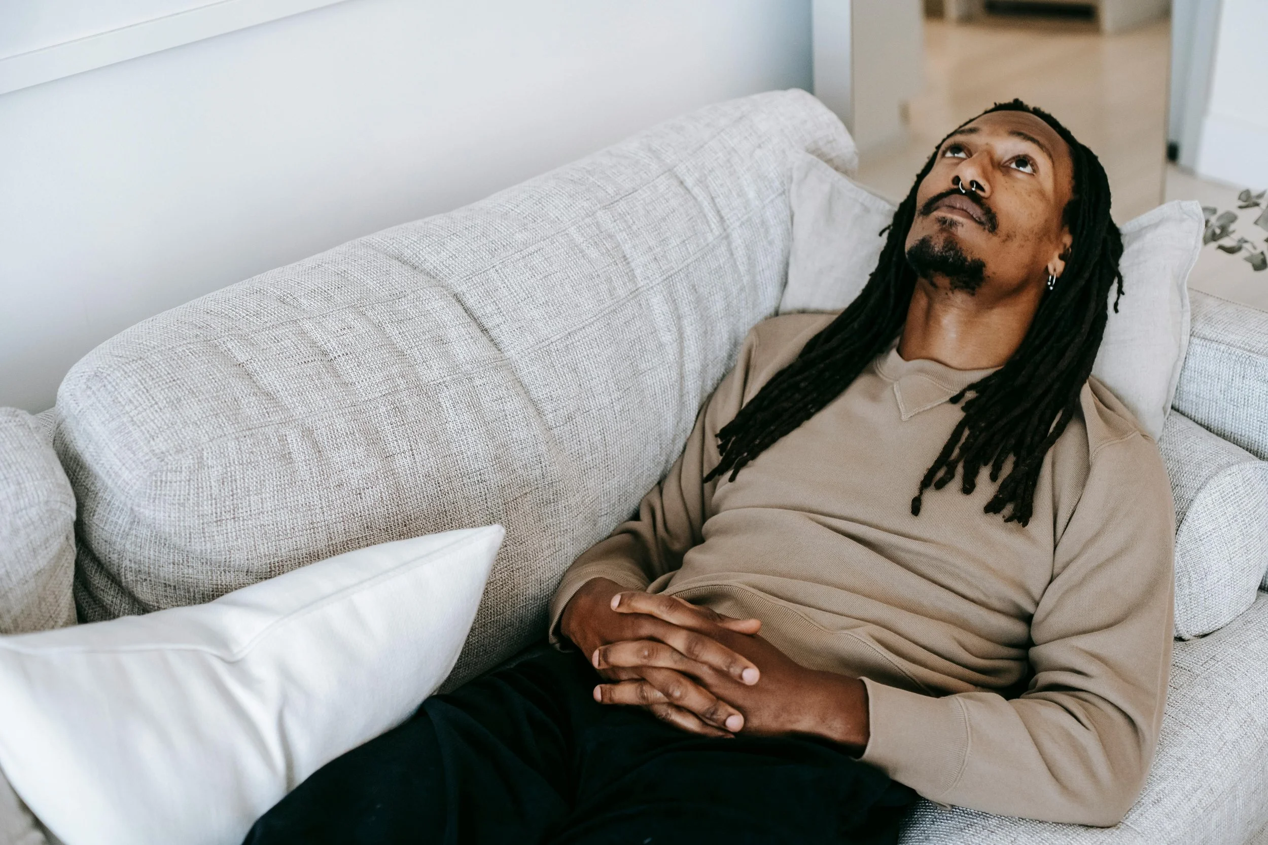 A man with dreadlocks and pierced nose lying on a beige couch, looking up.