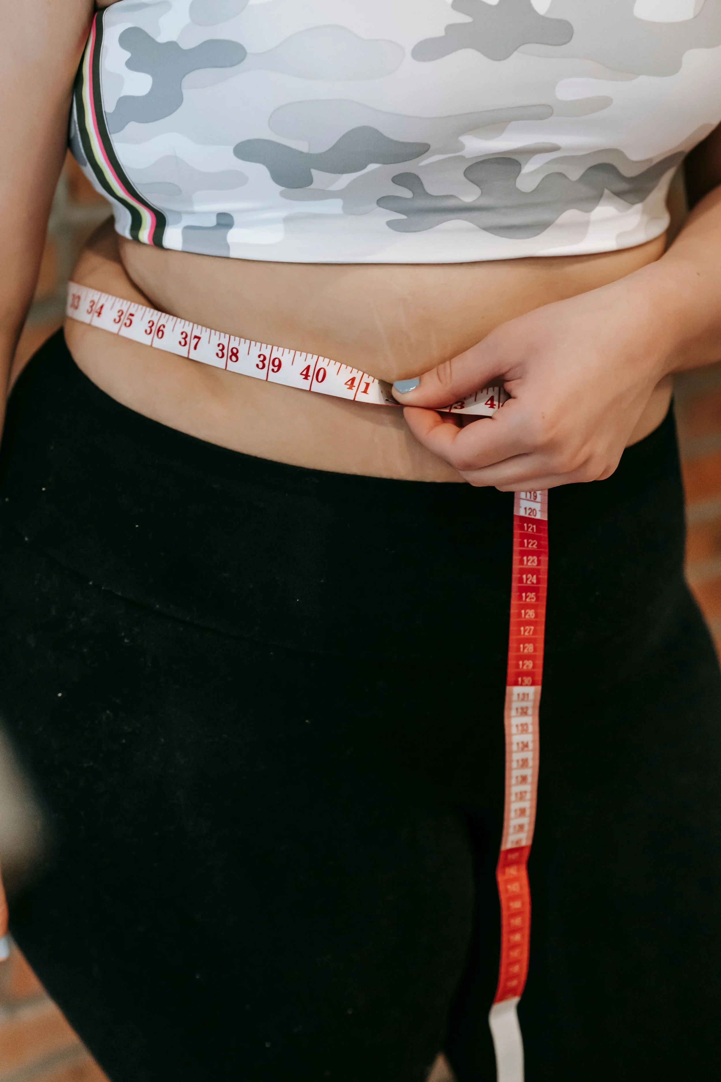 A person wearing a white camouflage tank top and black pants, measuring their waist with a red and white measuring tape.