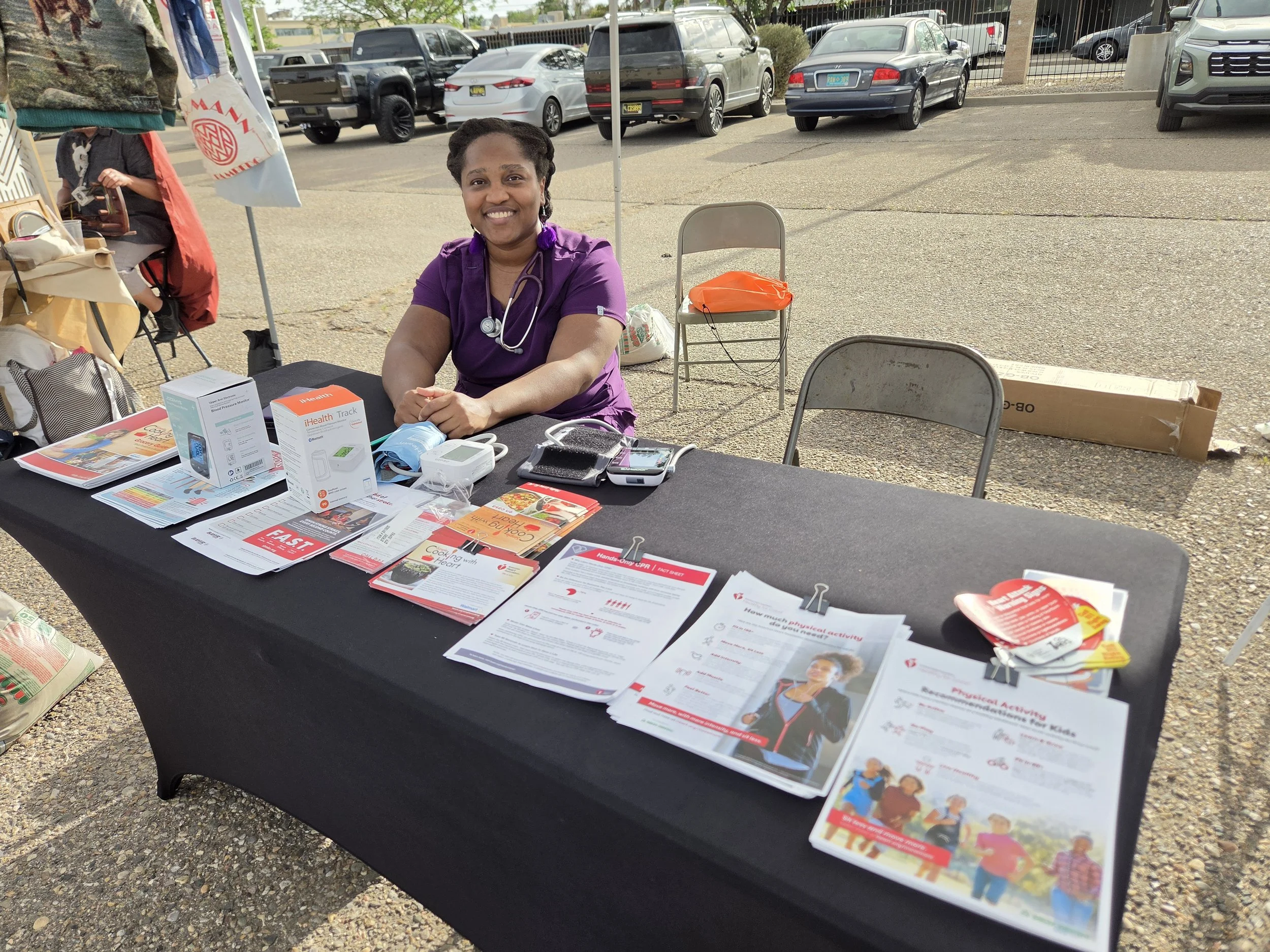 A woman in purple medical scrubs and a stethoscope smiling at a health awareness booth outdoors, with brochures and health-related materials on the table.