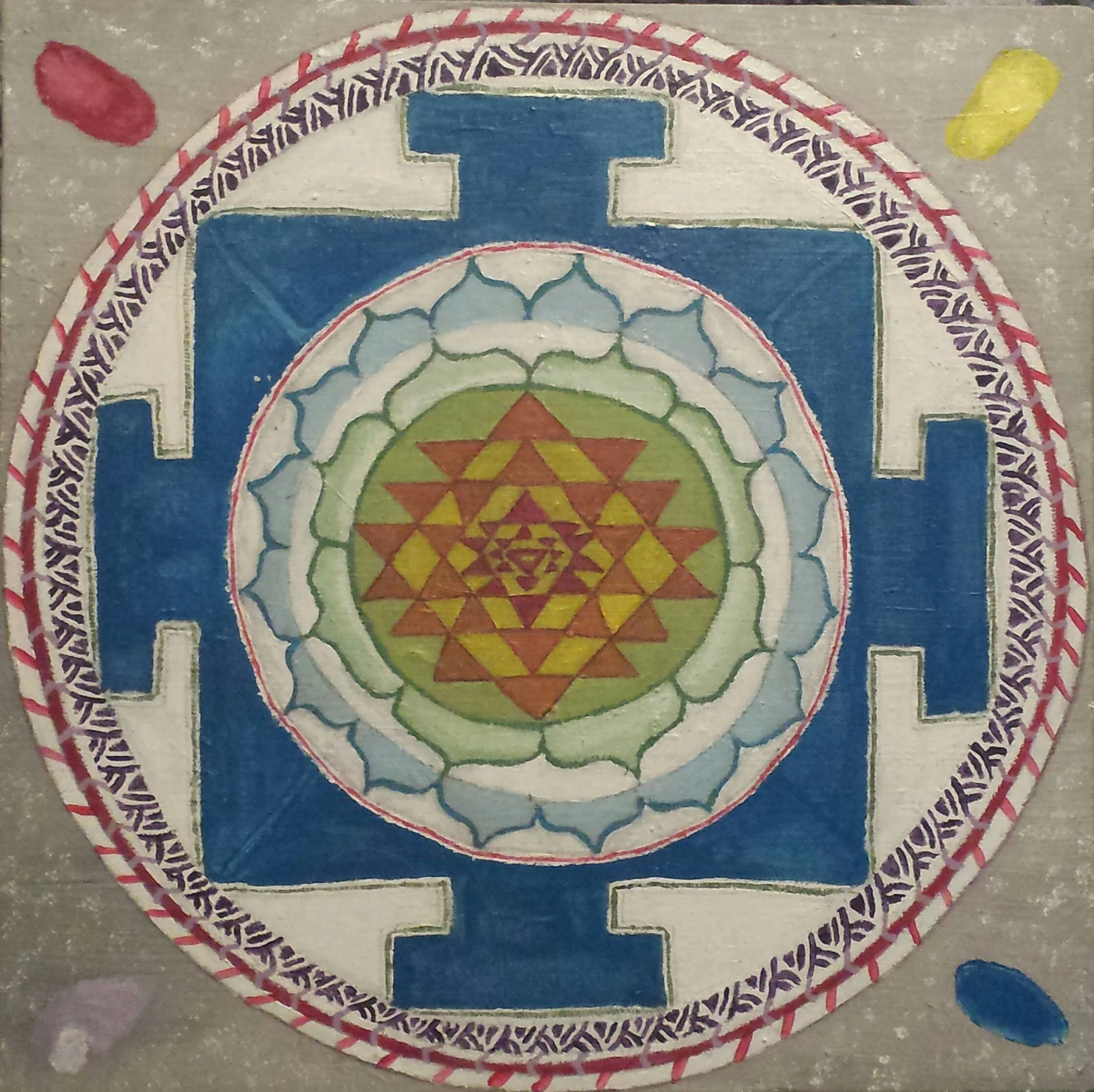Colorful circular rangoli design with geometric patterns, featuring a central yellow triangle, surrounded by green and red shapes, with a blue border, and decorative purple and white elements, on a gray floor with scattered colored powder.
