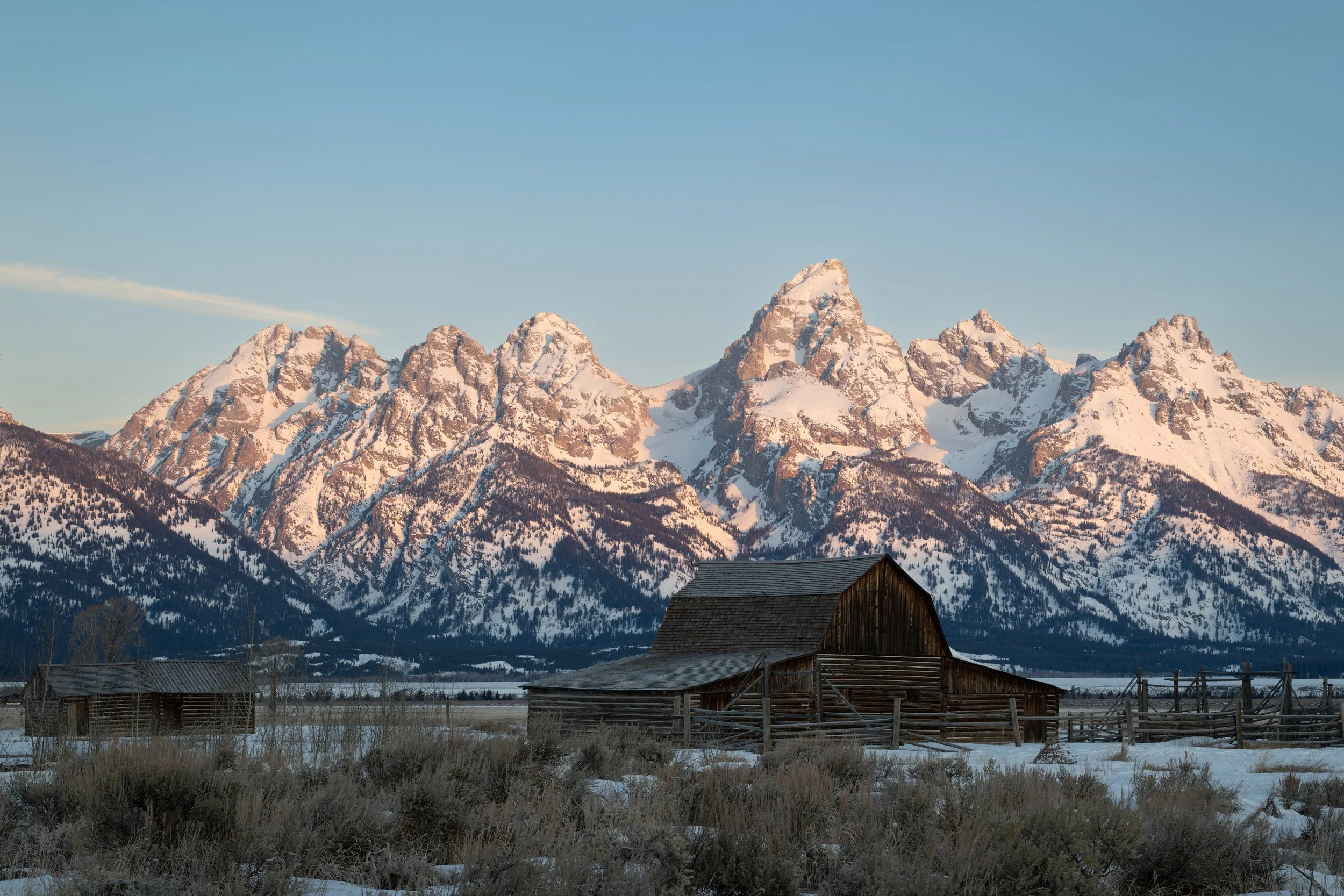 Snow-capped mountain range with a rustic wooden barn in the foreground and a clear blue sky.