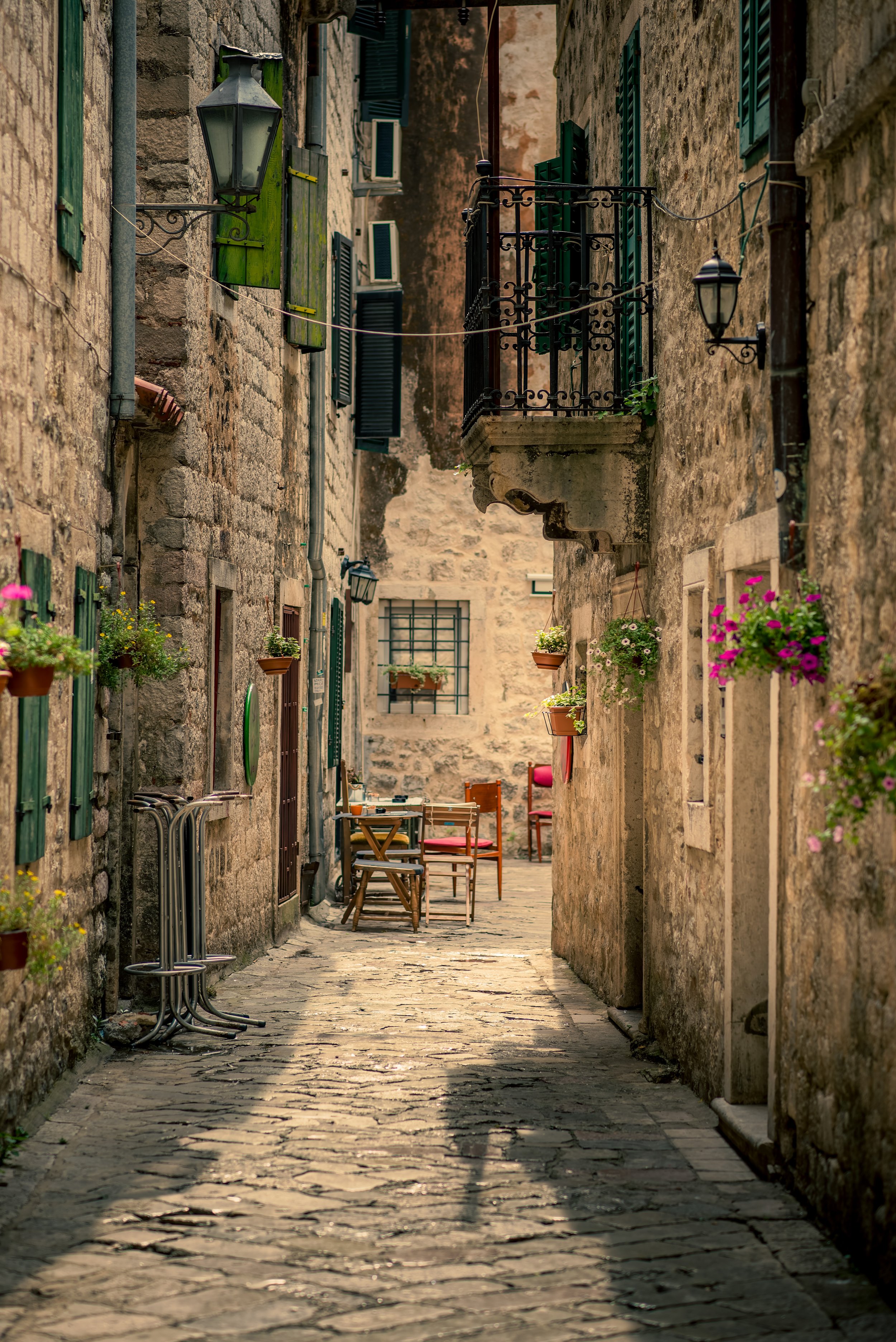 A narrow cobblestone street in an old European village, lined with stone buildings with green shutters and flower pots, with outdoor chairs and tables set up.