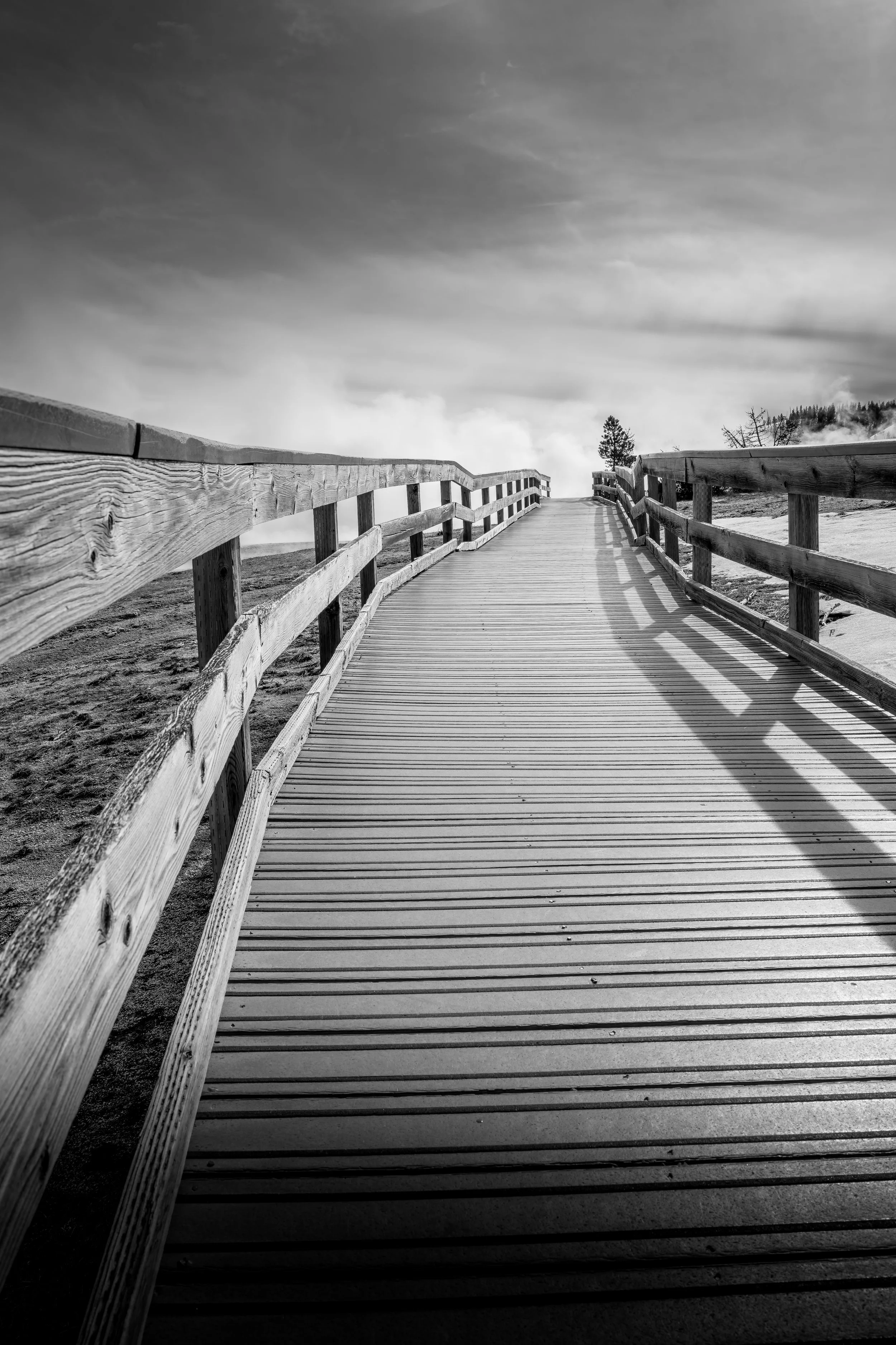 Wooden boardwalk stretching into the distance with guardrails on both sides, set in a natural landscape, under a cloudy sky in black and white. © 2026 Outgear Adventure Photography. Bruce Maxwell - Yellowstone NP