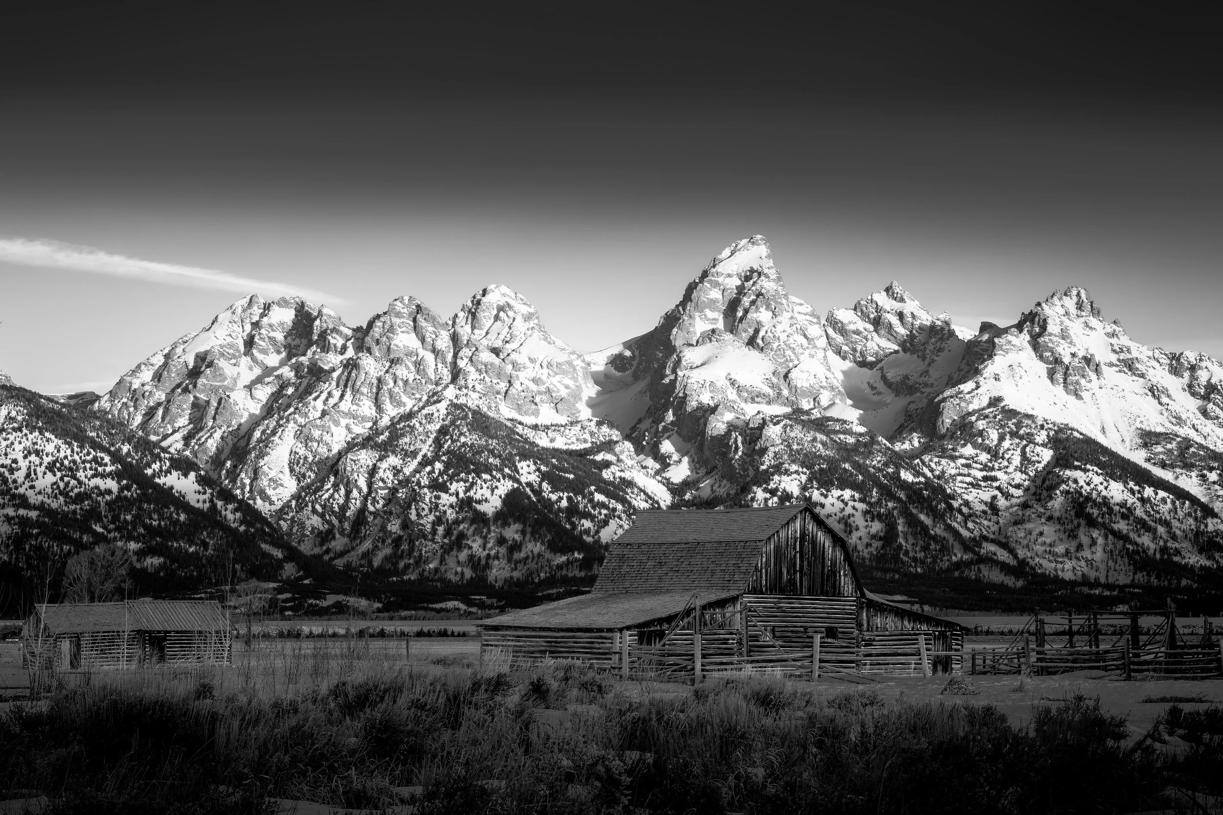 Snow-covered mountain peaks in the background with a wooden barn and fencing in the foreground, under a clear sky.