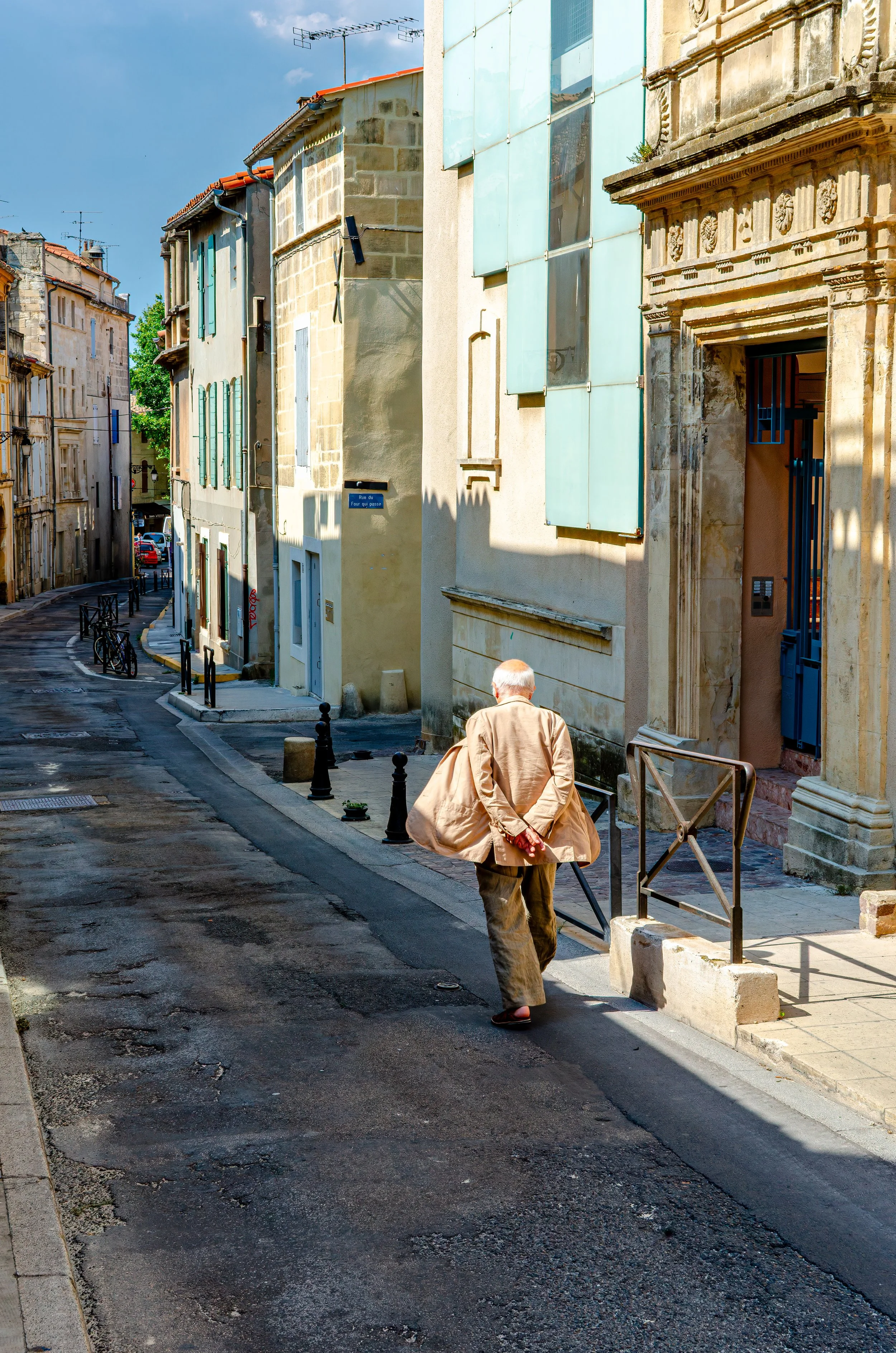 An elderly man with gray hair, wearing a beige coat, brown pants, and black shoes, walking down a narrow European street lined with old buildings with colorful shutters and ornate stone facades. © 2026 Outgear Adventure Photography. All rights reserv