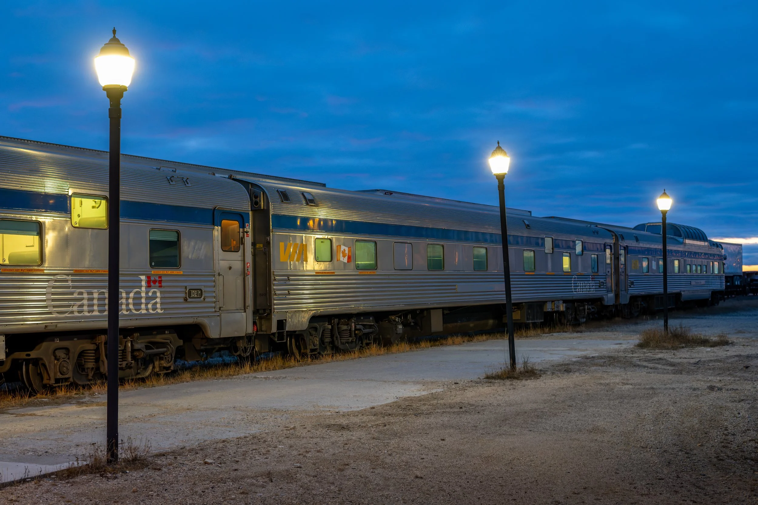 A silver Canadian train with lit windows parked outdoors at dusk, with three street lamps glowing. The sky is partly cloudy.