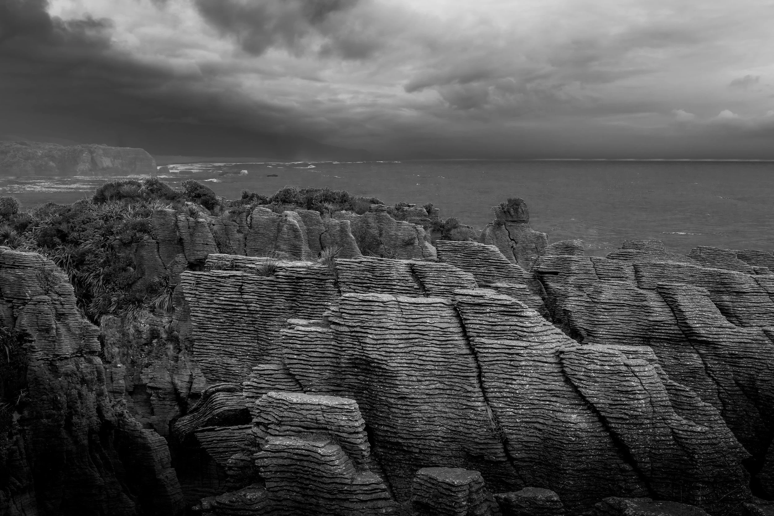 Cliffs and layered rock formations on a coastline with cloudy sky and ocean in black and white.