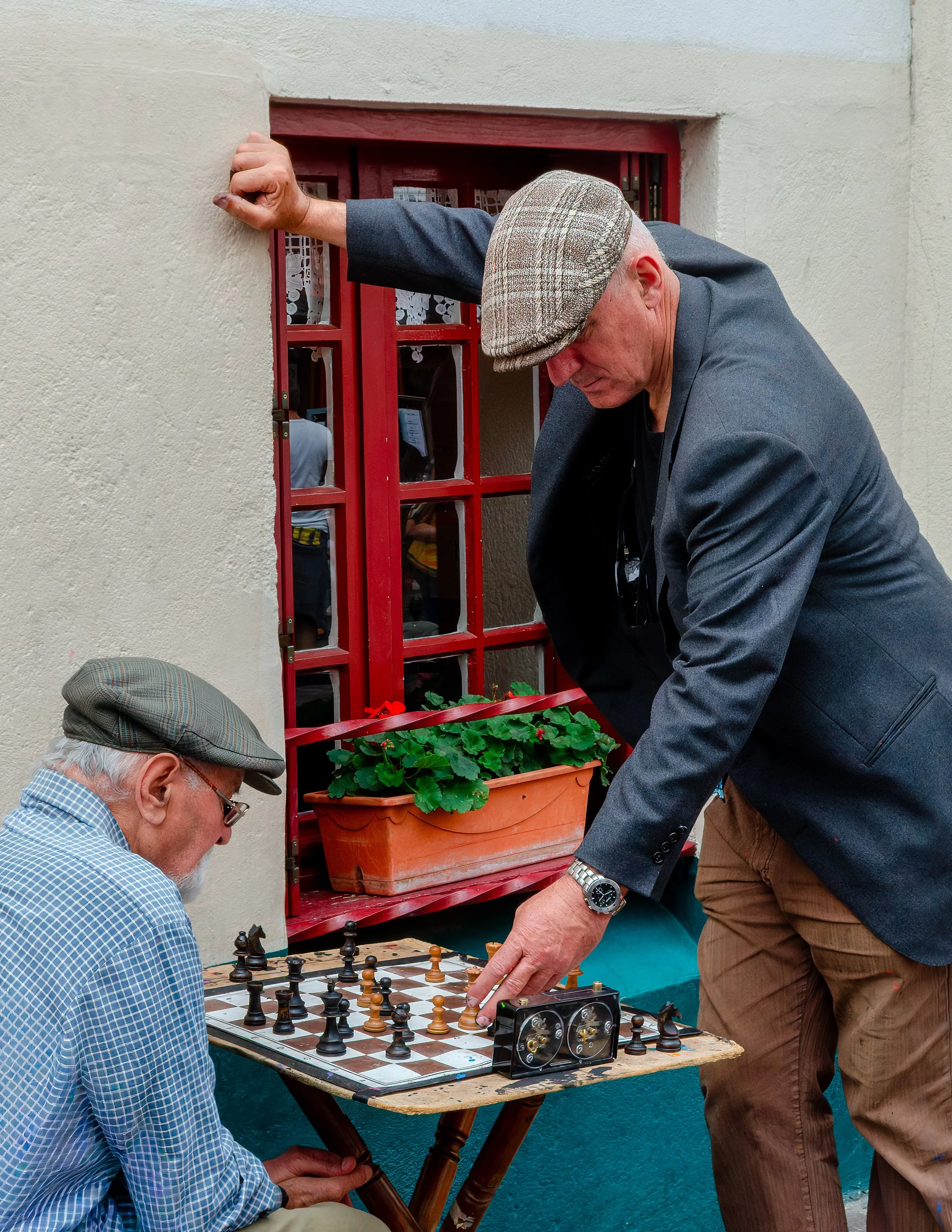 Two elderly men are playing chess outdoors. One man, with a white beard and glasses, is sitting and wearing a blue plaid shirt and a gray cap. The other man, who is standing and dressed in a dark blazer, beige pants, and a checkered cap, is moving a 
