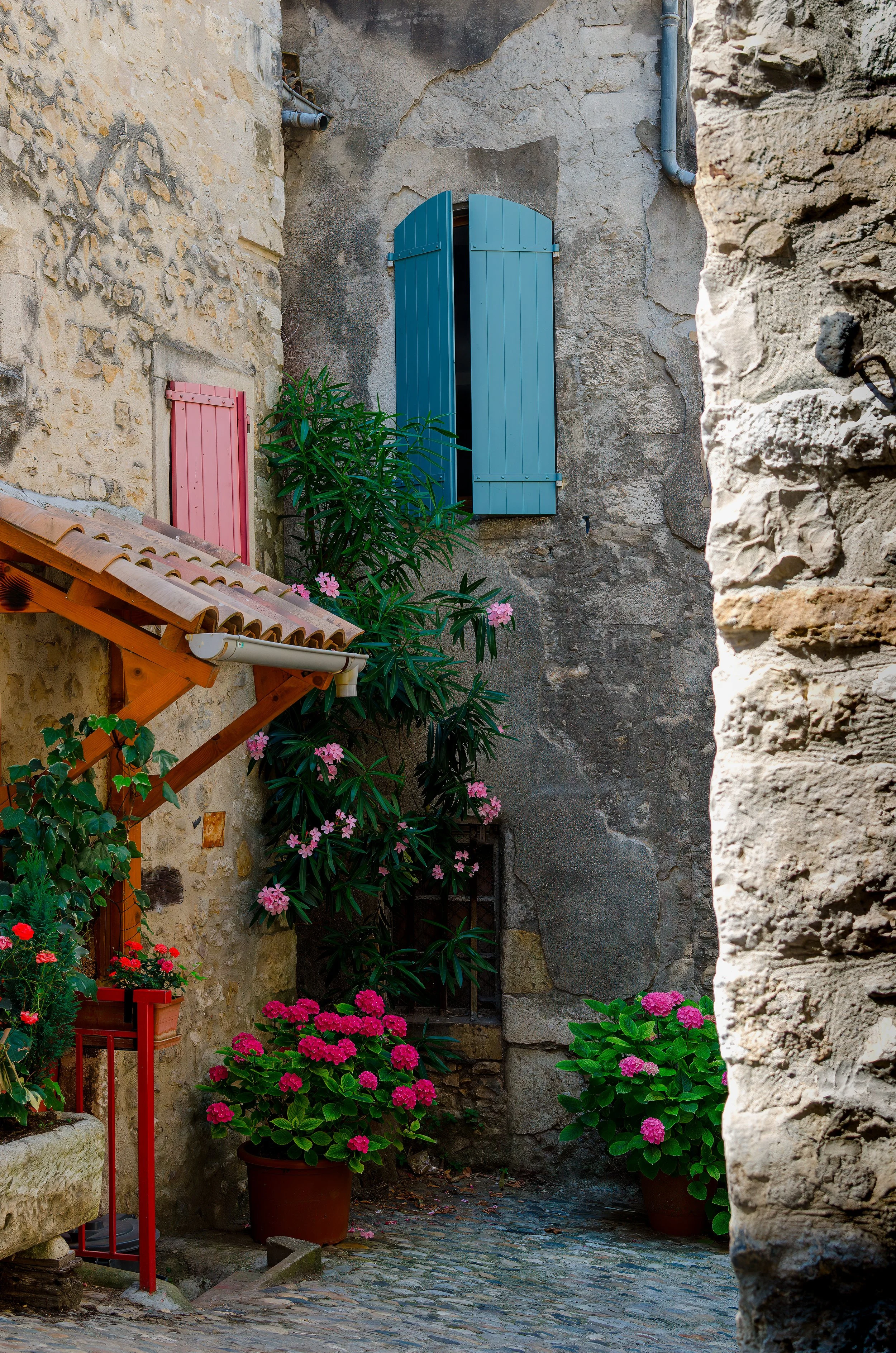 A narrow cobblestone alleyway in a historic European village, with stone walls, colorful flower pots, green plants, pink blossoms, and window shutters painted in blue and pink.