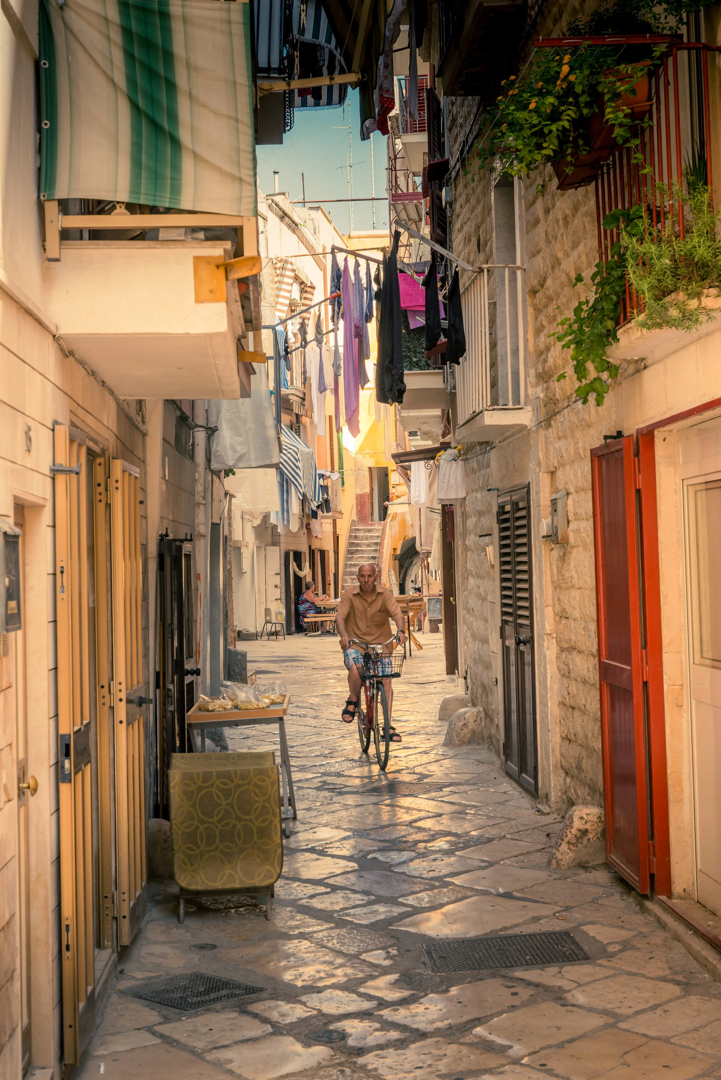 A narrow European street with stone paving, lined with shops and apartment buildings, a man riding a bicycle down the street, laundry hanging on lines above.