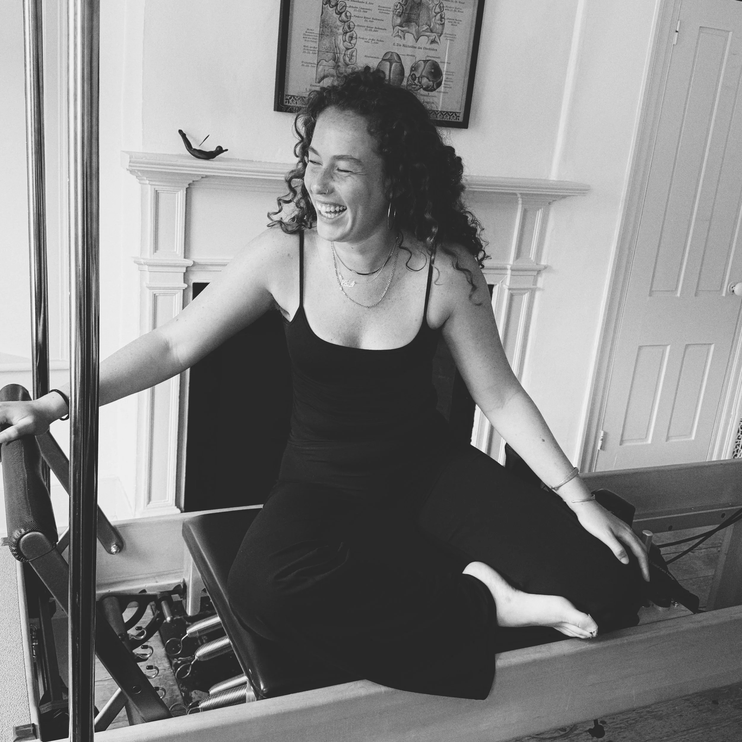 A woman with curly hair smiling and sitting on a chair in front of a mirror, wearing a black spaghetti strap top. The room has a white wall with a framed diagram of human teeth and a small sculpture of a reclining figure on a fireplace mantel. There are exercise springs and equipment in the foreground.