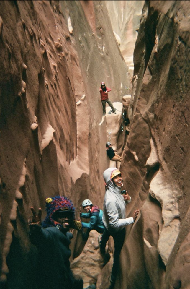 People exploring a narrow slot canyon with tall, smooth, reddish-brown walls, some climbing and others smiling and posing.